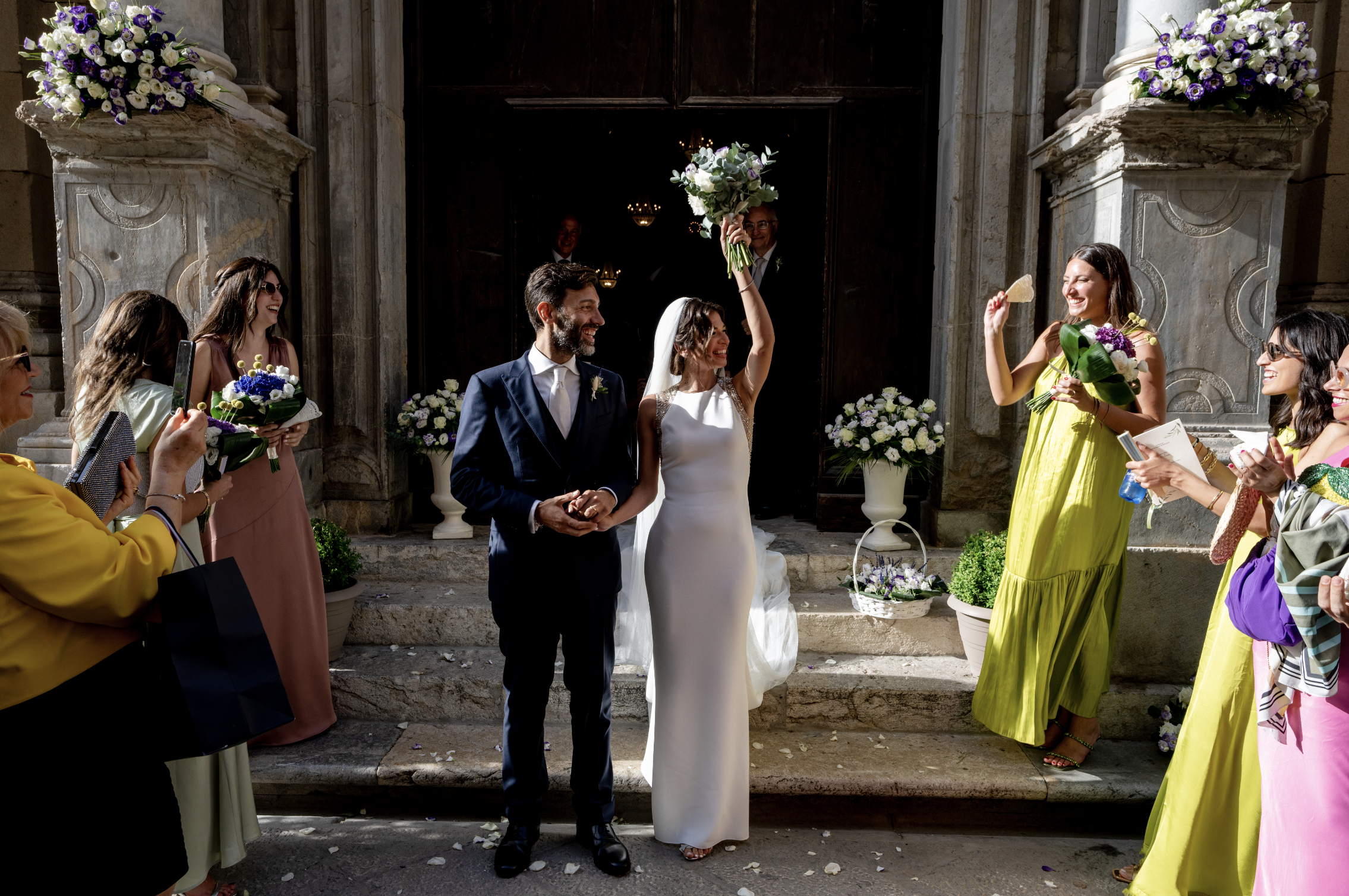 Bride and groom celebrating outside a Sicilian church surrounded by guests and elegant purple and white wedding florals.