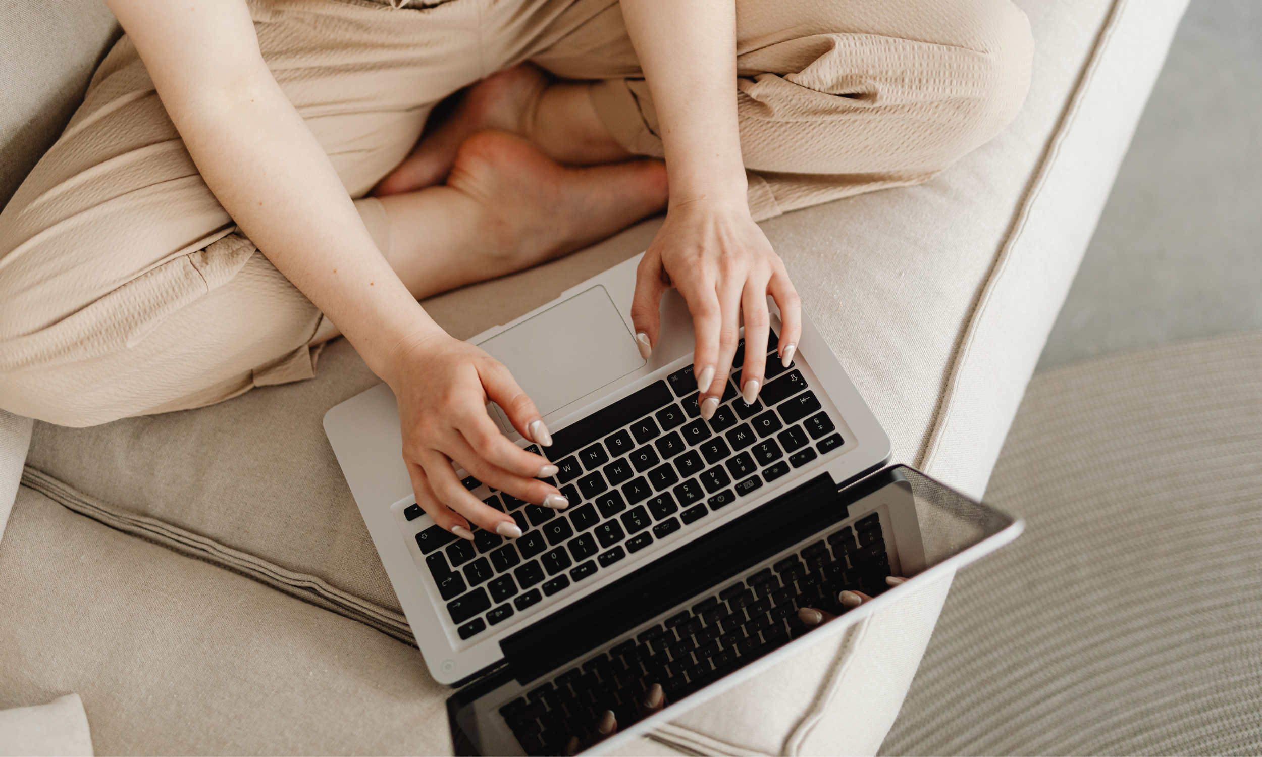 Woman sitting cross-legged on a beige couch, casually typing on a laptop placed in her lap, relaxed home setting.