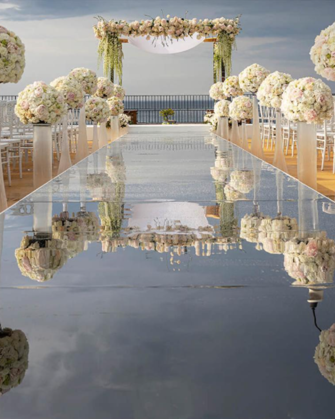 Luxury outdoor wedding ceremony in Italy with a mirrored aisle, white floral arrangements, and a decorated arch overlooking the sea