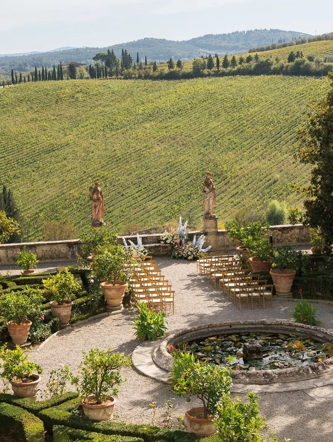 Romantic Italian garden wedding ceremony set on a stone terrace overlooking rolling vineyards, with gold chairs arranged around a floral ceremony space