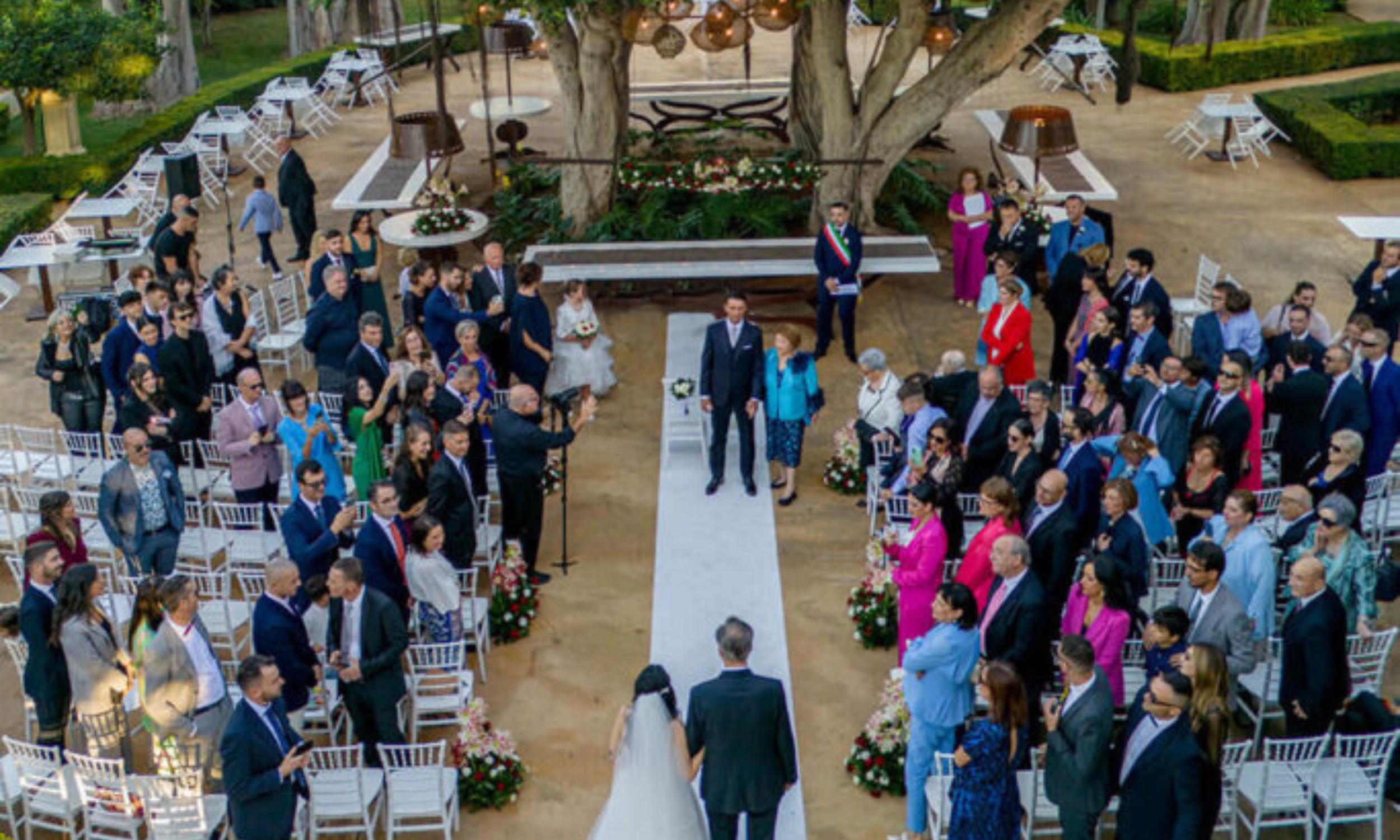 Outdoor wedding ceremony in an Italian garden with guests seated around a central aisle, bride walking toward the groom under large trees