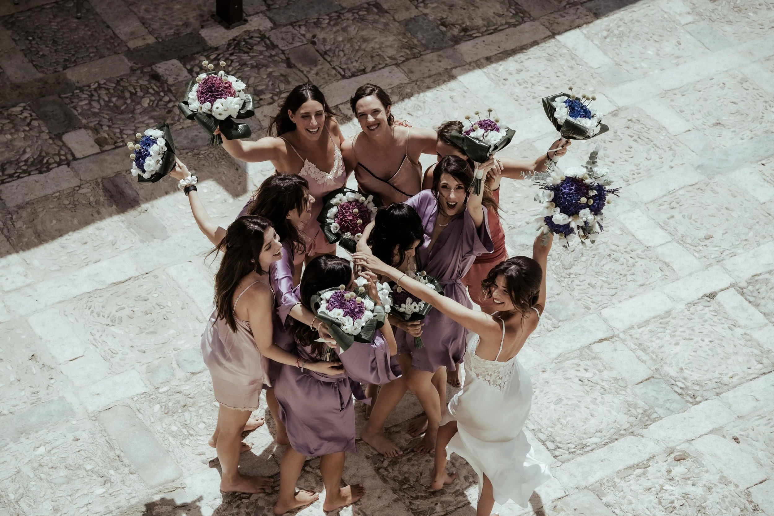 Bridesmaids gathered together holding purple and white wedding bouquets during a joyful Italian wedding celebration.