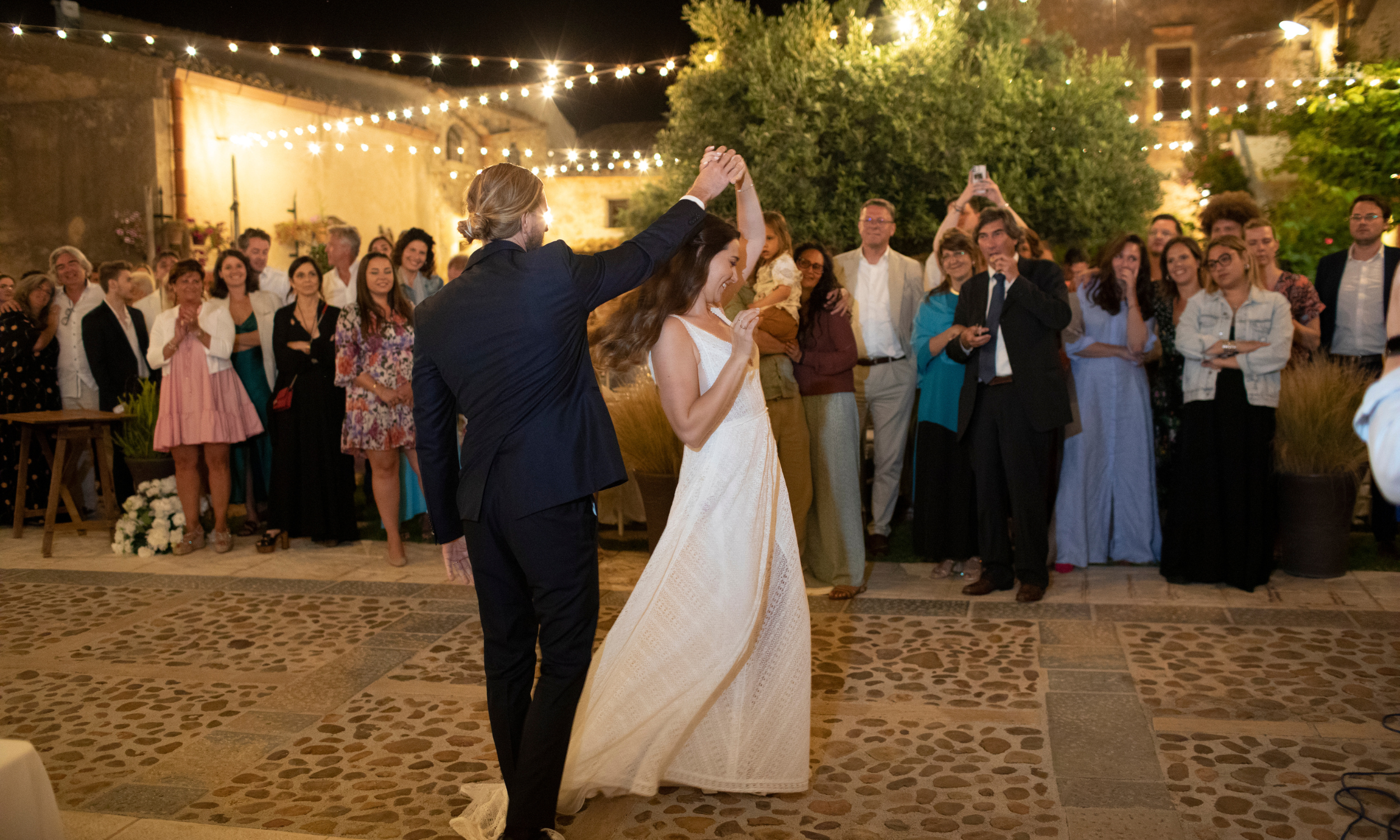 Bride and groom sharing their first dance under string lights in a rustic Sicilian courtyard, surrounded by smiling guests in evening attire.