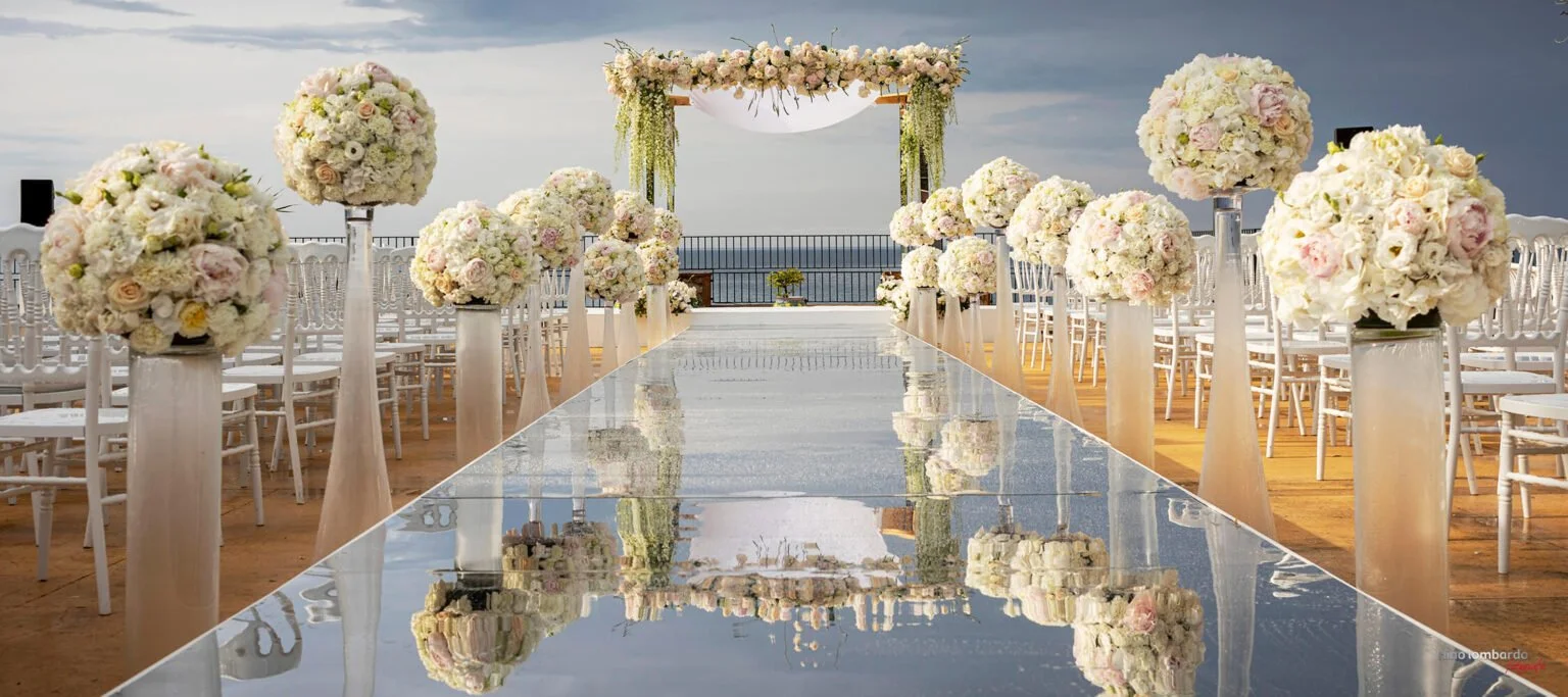 Luxury seaside wedding ceremony setup in Italy with white floral arrangements, mirrored aisle, and ocean views.