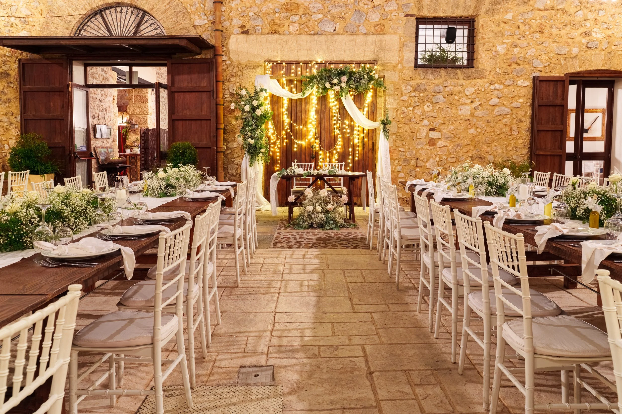 A warmly lit indoor wedding ceremony aisle inside a rustic stone Italian venue, with white chiavari chairs, fairy lights draped overhead, and a floral arch framing the entrance.