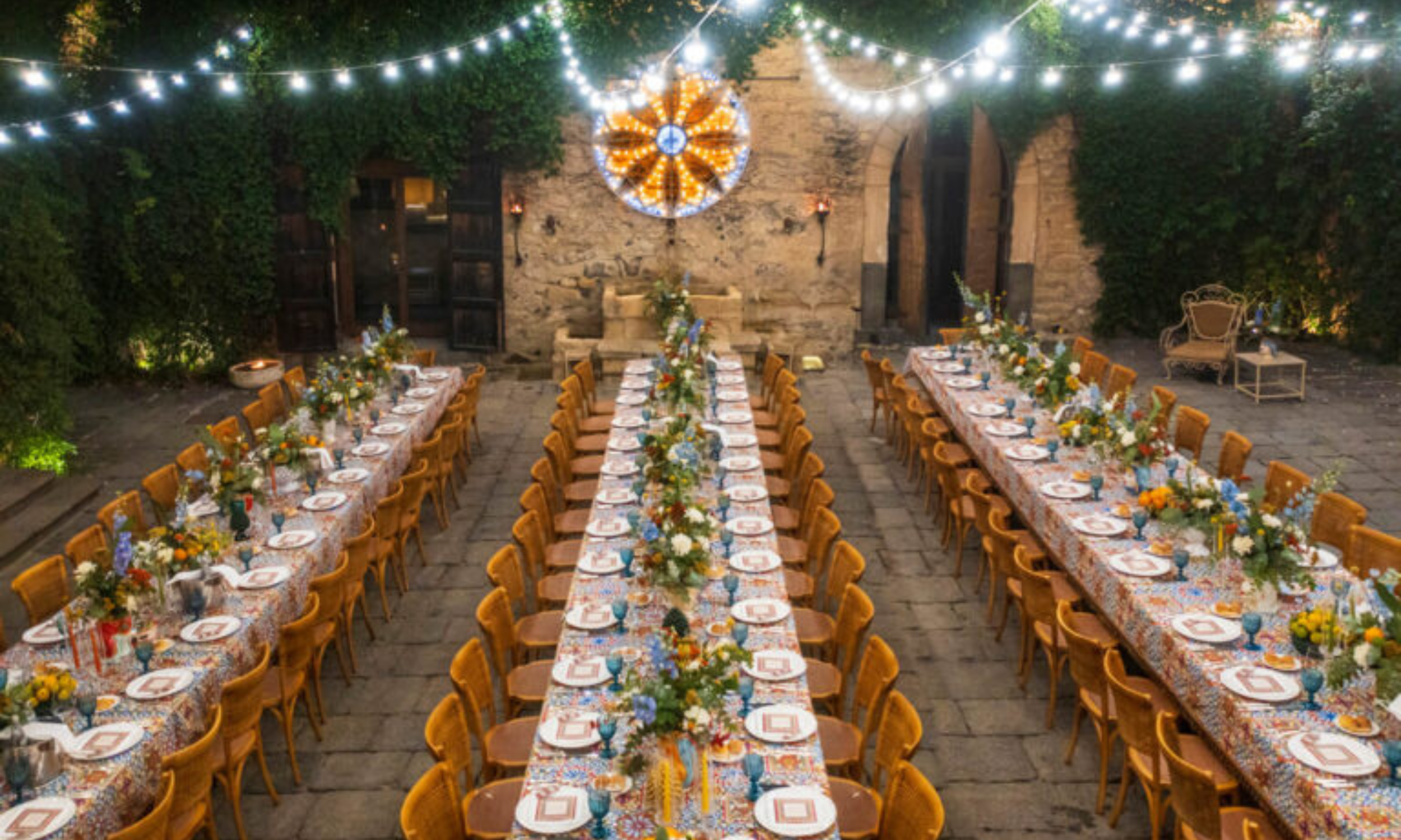 Long table wedding reception in an Italian courtyard with colourful table settings, floral arrangements, and string lights overhead at night