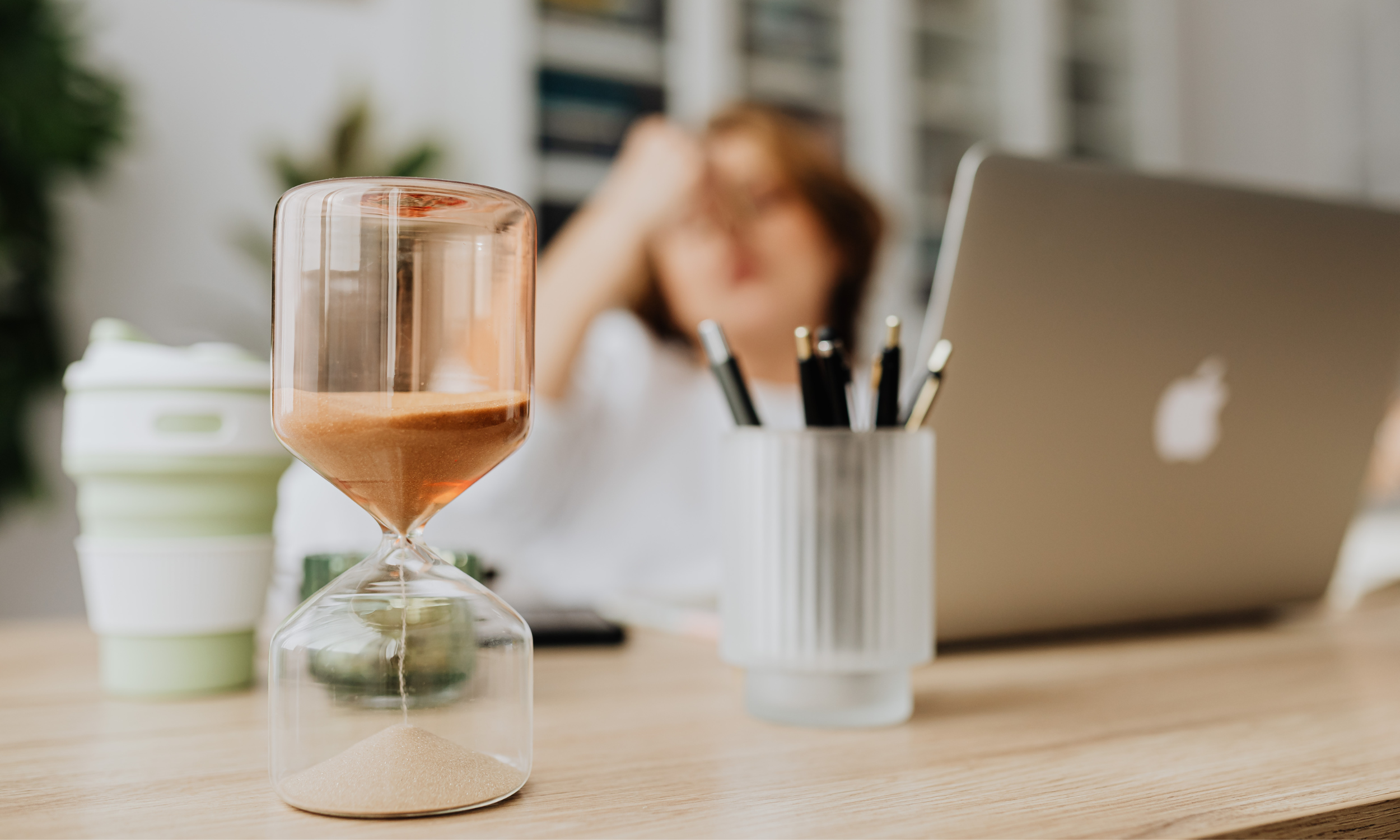Clear hourglass with brown sand on a desk, with a laptop and pens in the background and a frustrated woman slightly blurred in the distance.