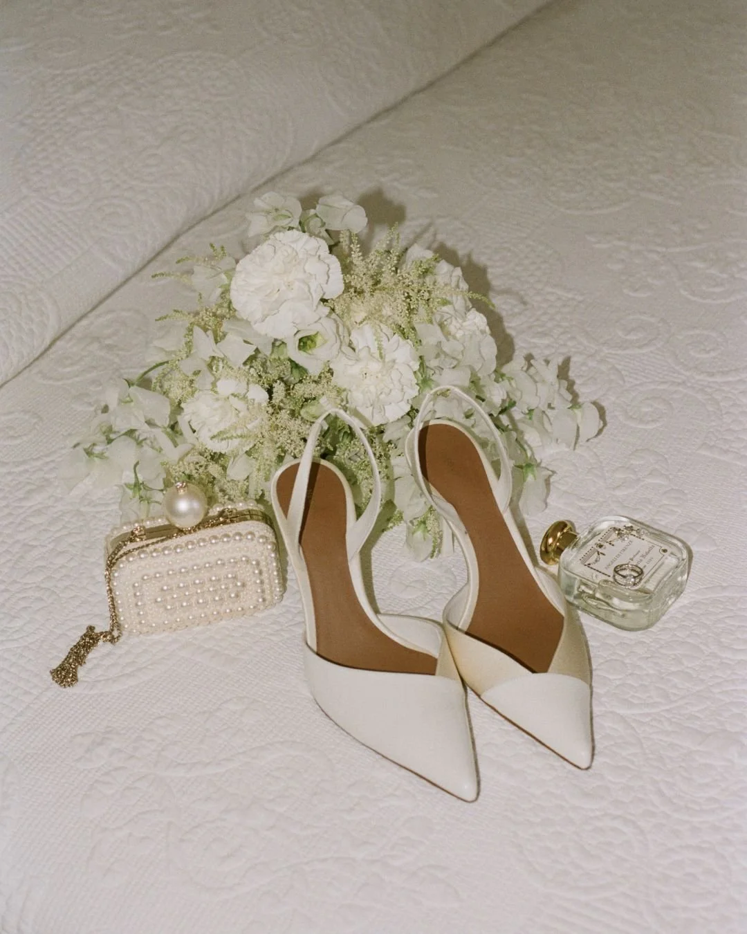 White bridal shoes, pearl handbag, perfume and white wedding bouquet arranged on a bed.