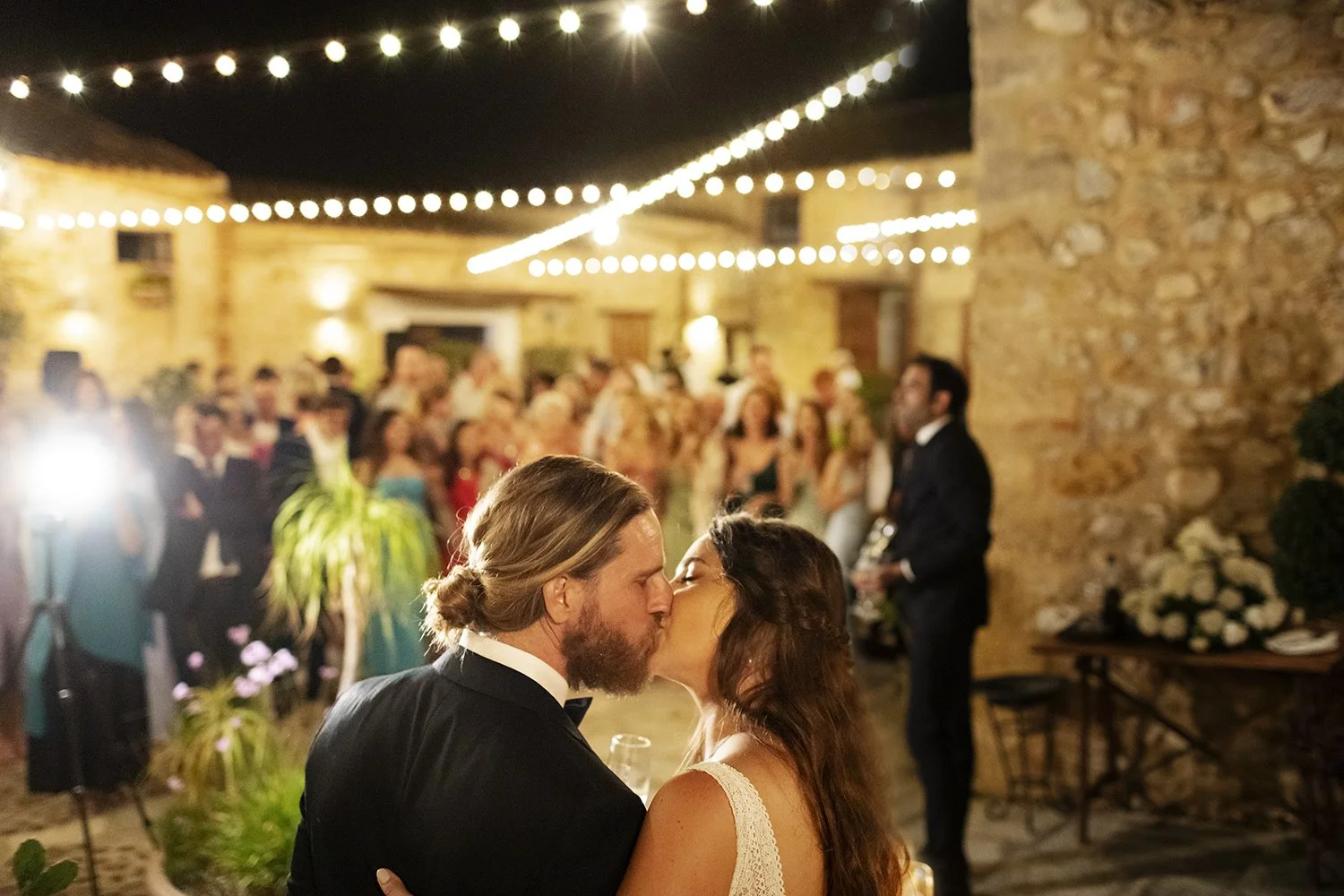Bride and groom sharing a romantic first dance kiss under glowing string lights in Baglio Aversa’s rustic stone courtyard, surrounded by wedding guests at night.