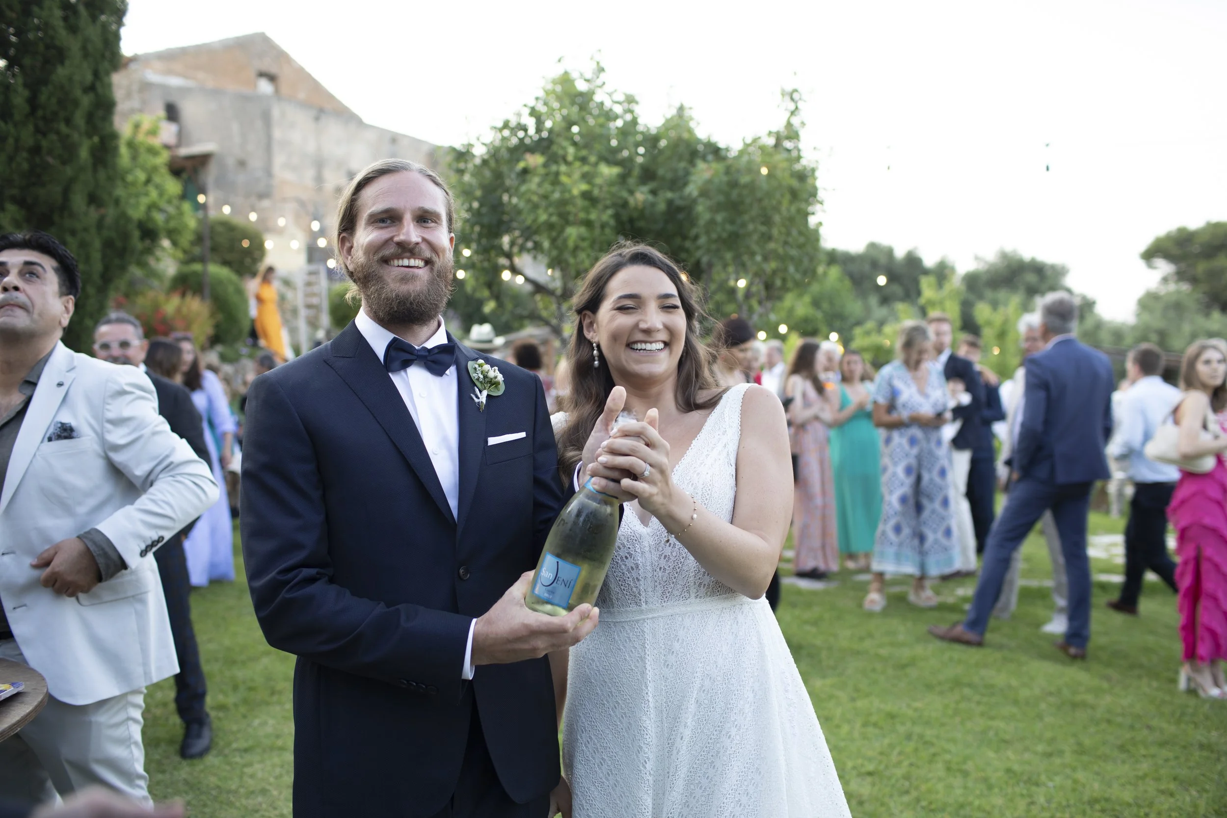 A smiling bride and groom laughing together as the groom holds a champagne bottle at an outdoor wedding reception with guests celebrating in the background.