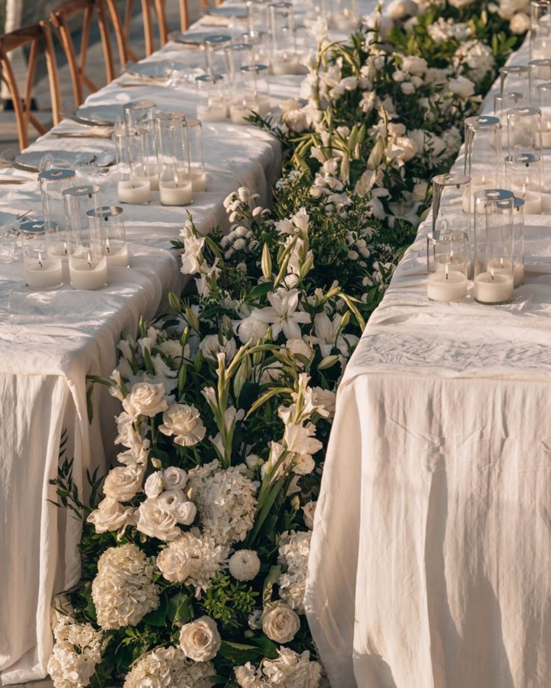 Elegant outdoor wedding reception tablescape with white linen, glass candle holders, and a lush white floral runner between long banquet tables.
