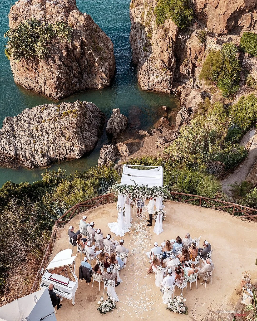 Aerial view of a cliffside wedding ceremony in Sicily overlooking the sea with guests and floral setup
