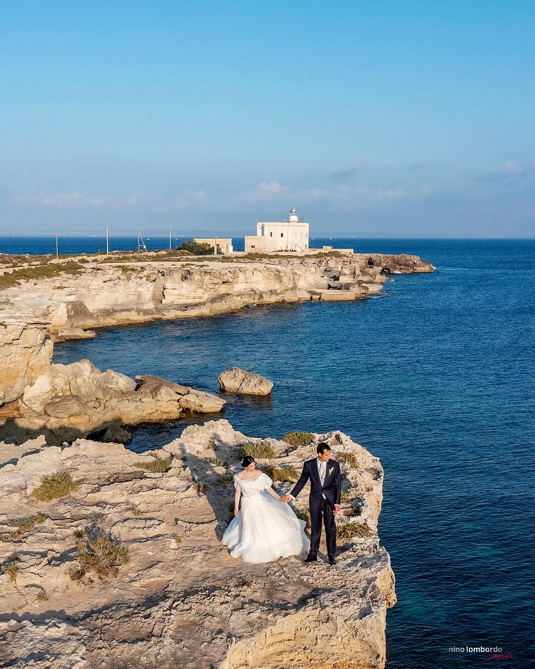 Bride and groom standing on a rocky cliffside overlooking the sea in Sicily with lighthouse and coastal views