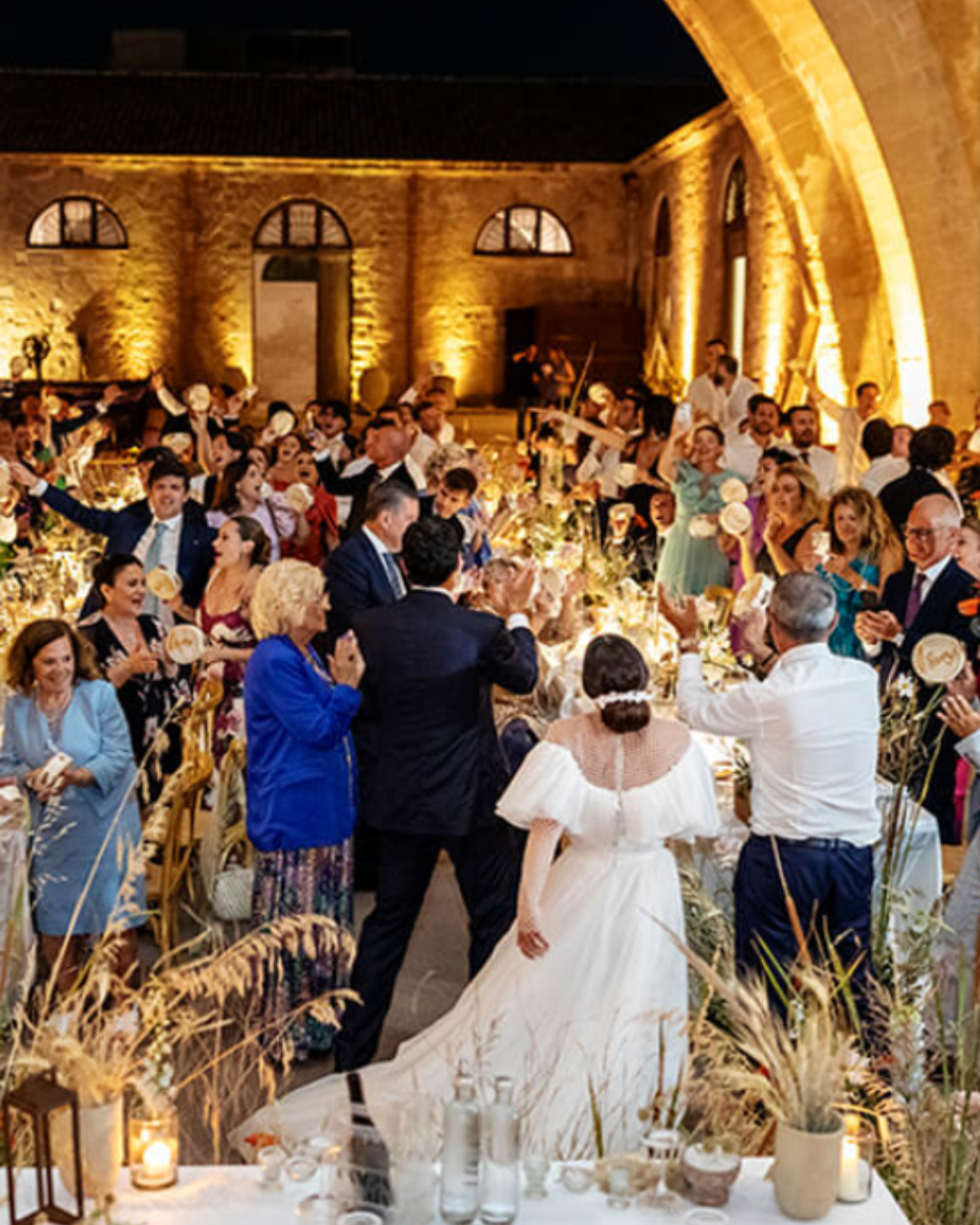 Italian wedding reception at night in a rustic courtyard with guests celebrating, warm lighting, long tables, and the bride and groom walking through the crowd