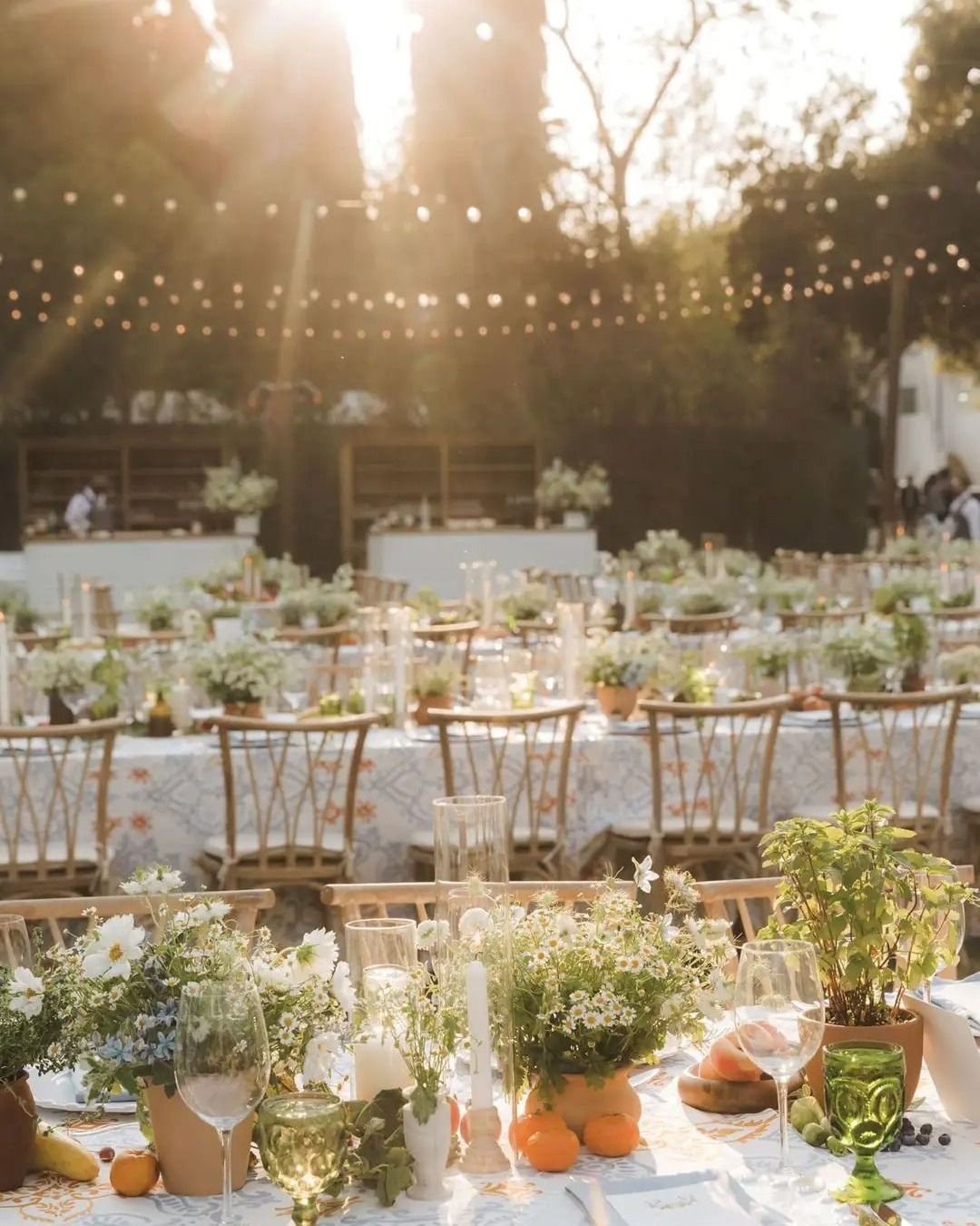 Outdoor wedding reception at golden hour with long tables, floral centrepieces, string lights overhead, and place settings in the foreground.