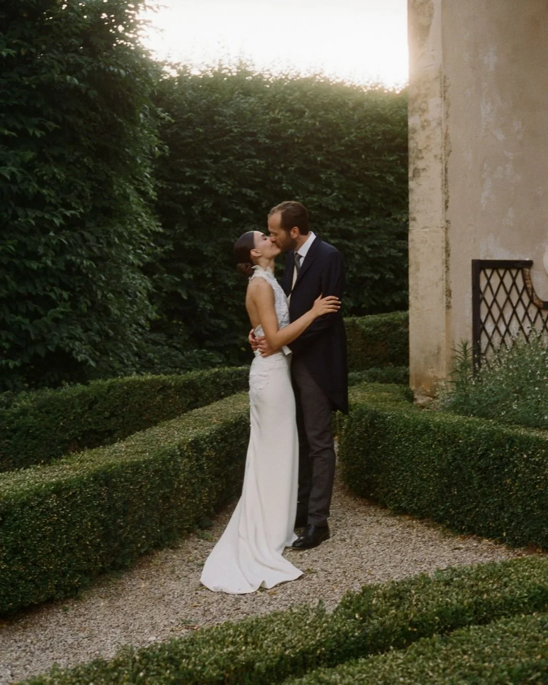 Bride and groom kissing among manicured gardens beside a historic Italian villa.