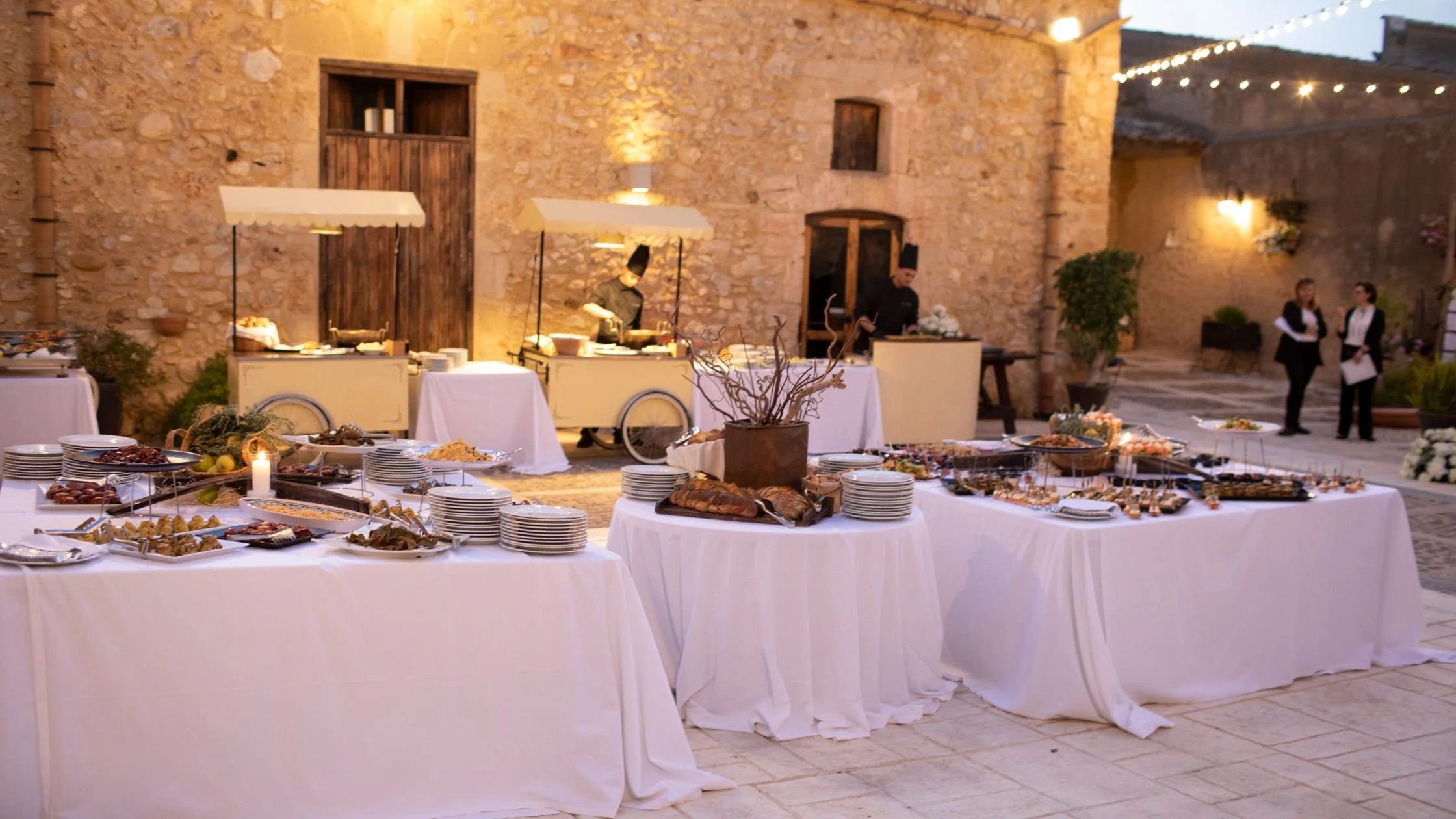 Rustic Sicilian wedding reception with buffet tables set under string lights in a historic stone courtyard.