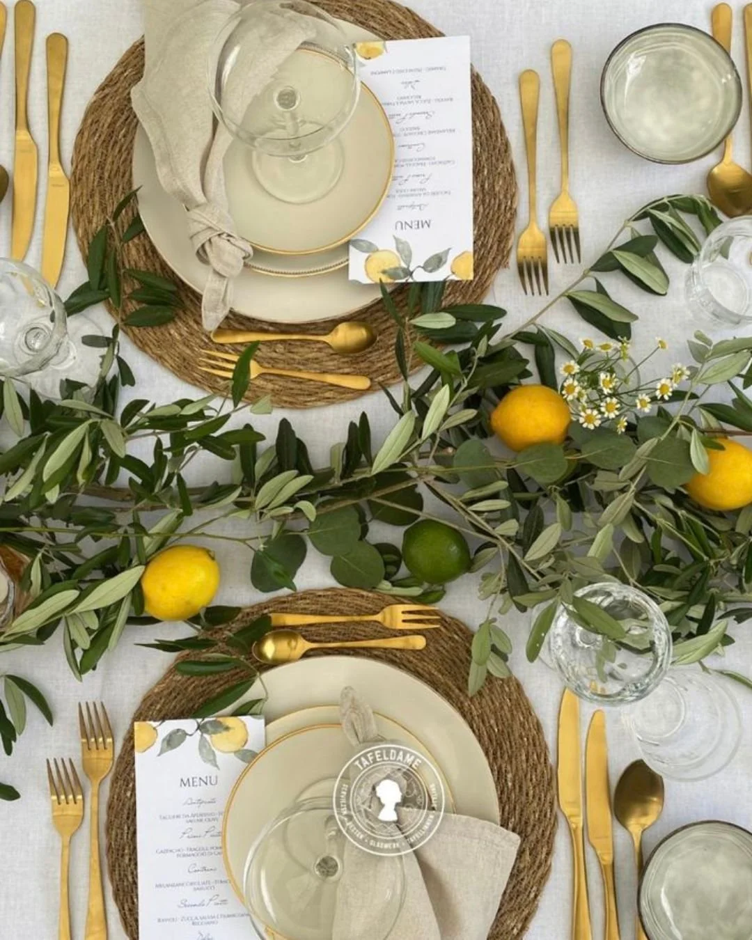 Overhead wedding tablescape with lemon-accented menus, gold cutlery, linen napkins, and olive branches with lemons on a white tablecloth.