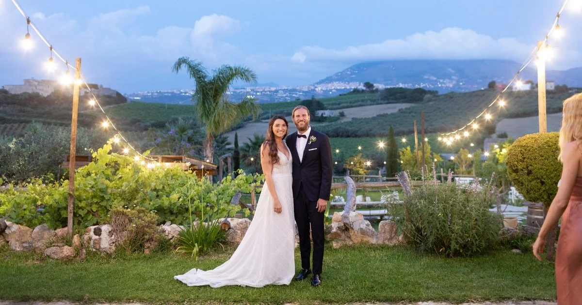 Bride and groom standing in a vineyard at sunset during their wedding in Italy, with string lights and rolling hills in the background.