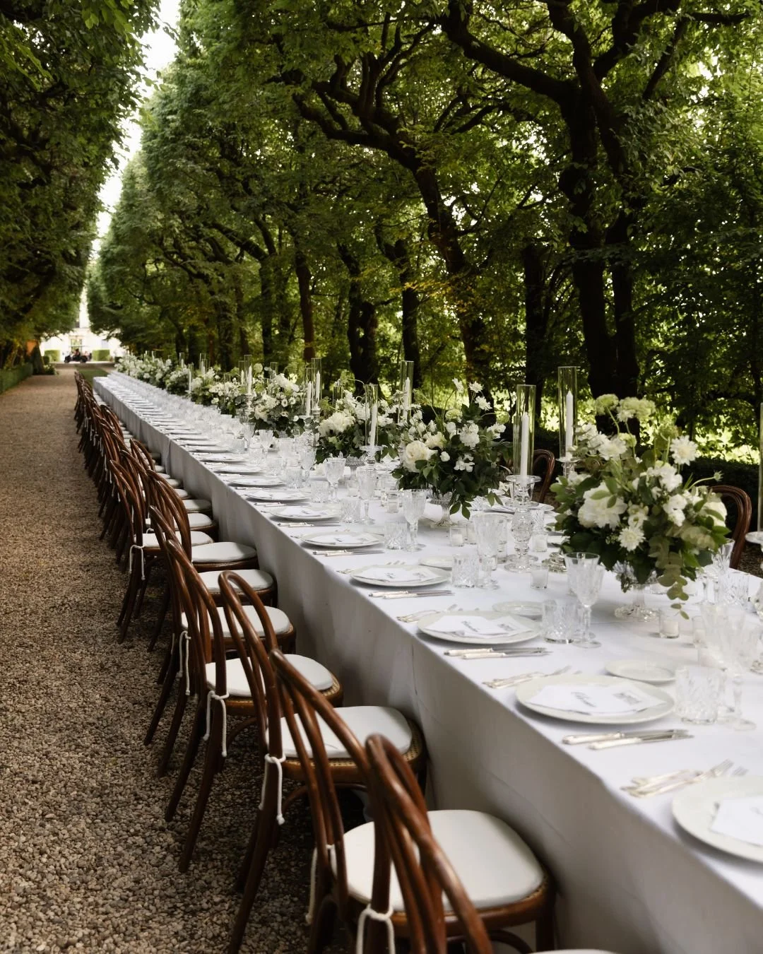 Long outdoor wedding reception table with white flowers, candles and wooden chairs beneath lush Italian trees.