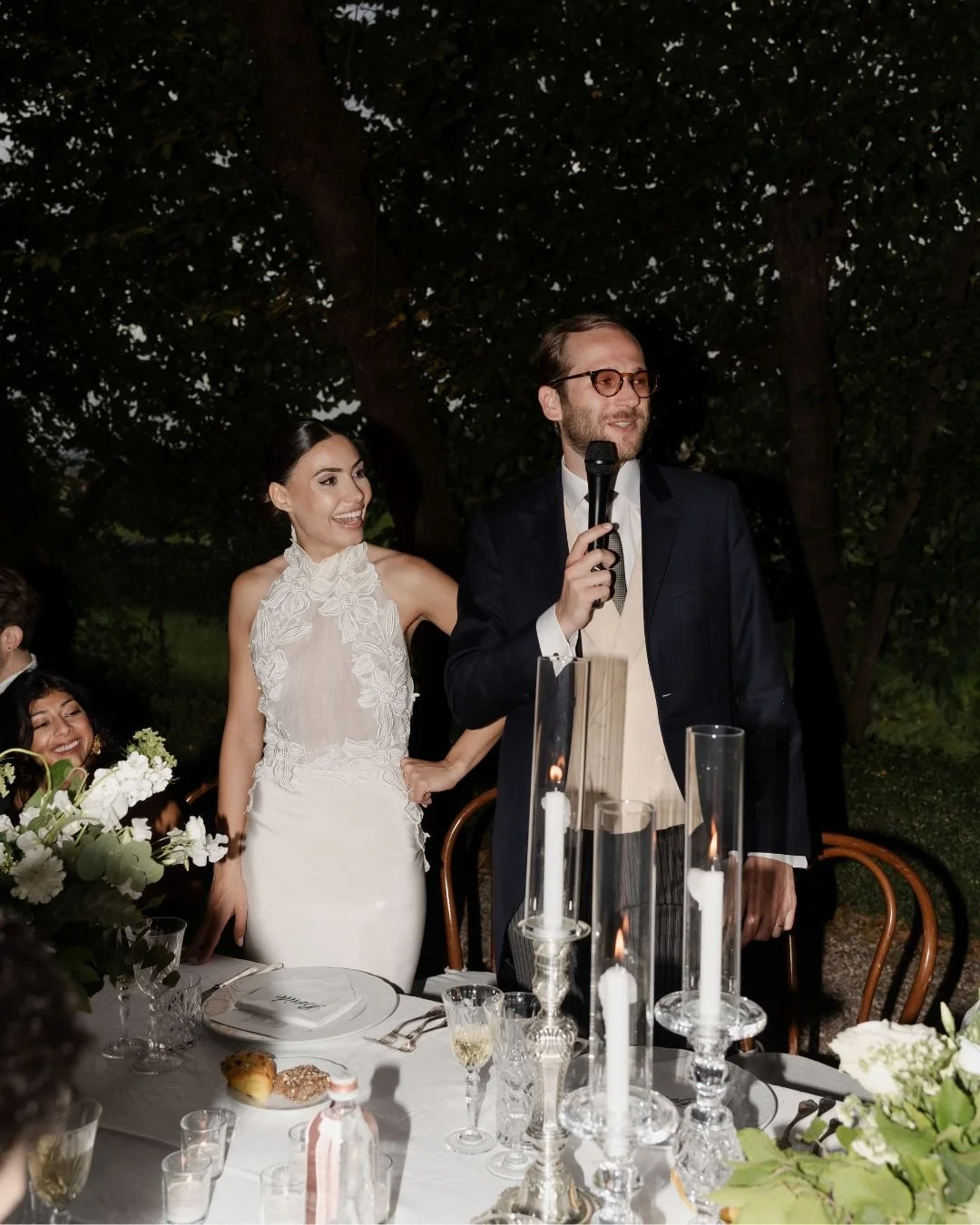 Bride smiling beside the groom as he gives a speech during an elegant outdoor wedding reception in Italy.