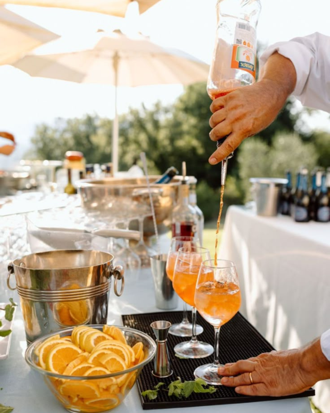 Bartender pouring Aperol into glasses for spritz cocktails at an outdoor Italian wedding, with sliced oranges and prosecco bottles on a sunlit bar setup.