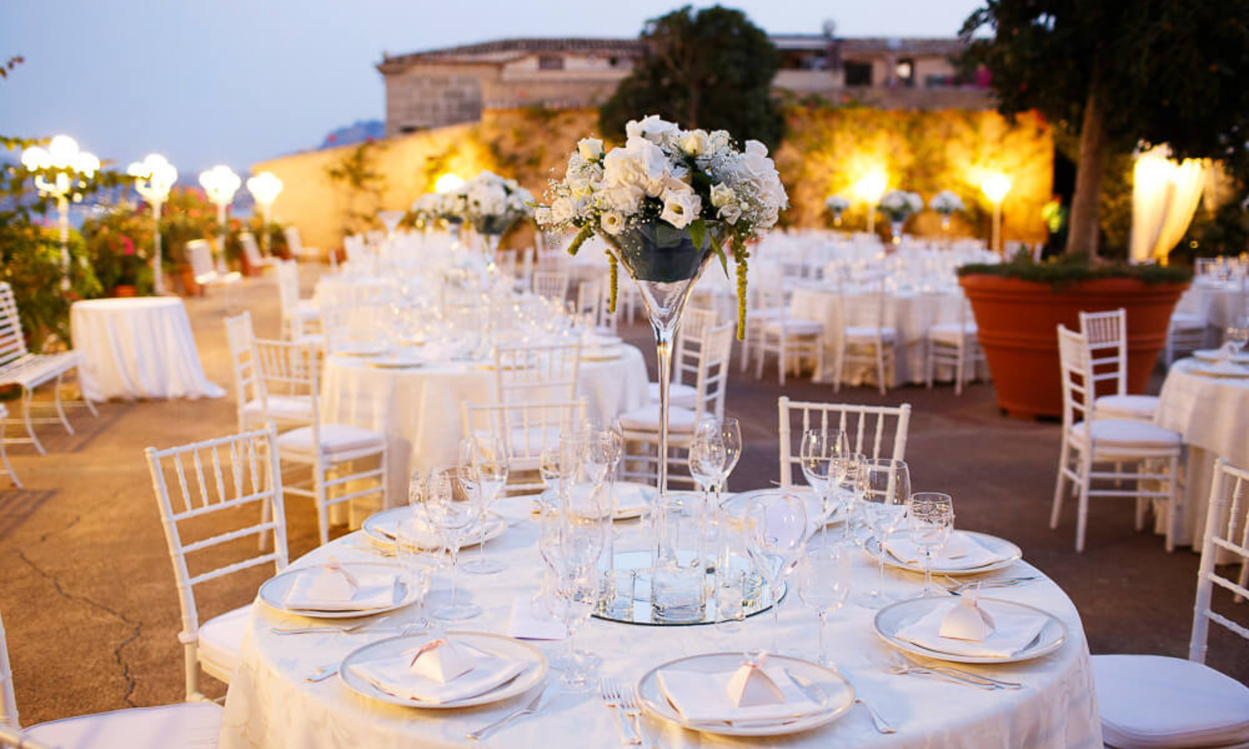 Elegant outdoor wedding reception table setting in Italy with white linens, glassware, and floral centrepiece at sunset