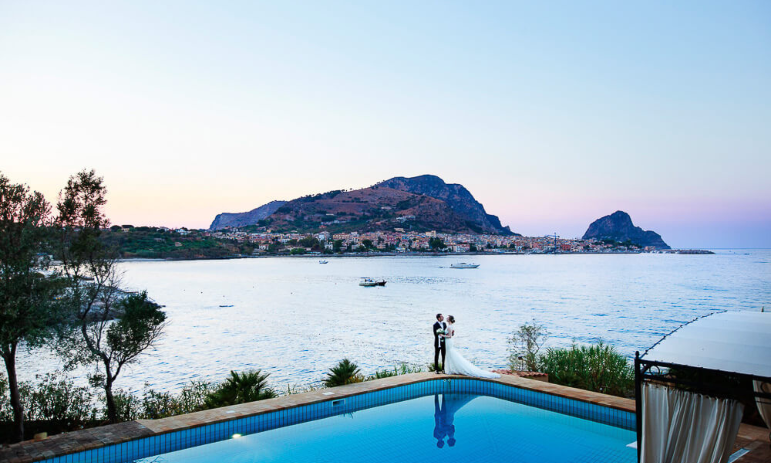 Bride and groom standing by a pool overlooking the sea in Sicily at sunset with coastal town and mountains in the background