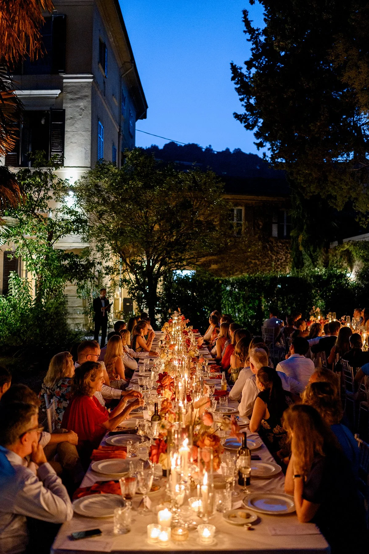 Guests seated at a long candlelit wedding reception table in the garden of an Italian villa at dusk, surrounded by elegant floral arrangements and warm ambient lighting.