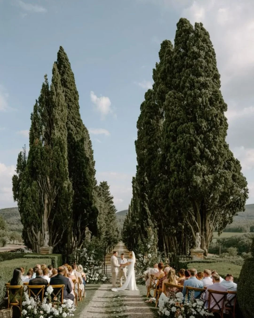 Outdoor wedding ceremony in Tuscany with a couple exchanging vows beneath tall cypress trees and guests seated along a garden aisle.