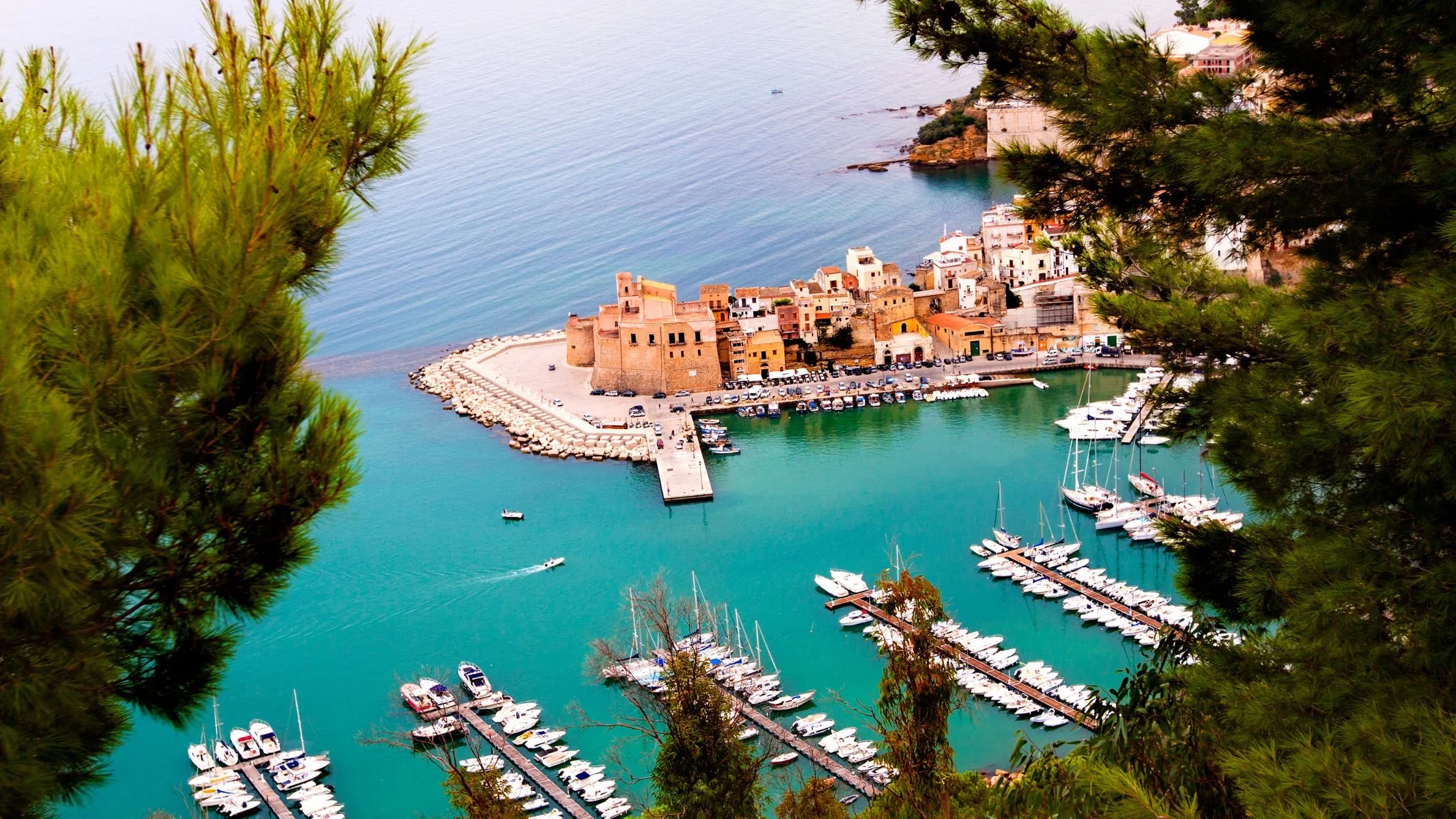 Scenic view of Castellammare del Golfo harbor in Sicily, with a historic castle overlooking turquoise waters and a marina filled with boats.