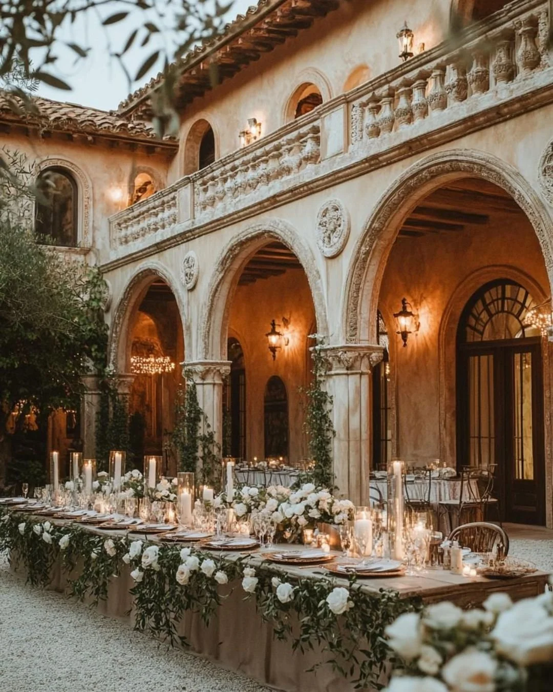 Romantic Italian courtyard wedding reception with long banquet table, white florals and candles, set beneath warm-lit stone arches and a balcony.