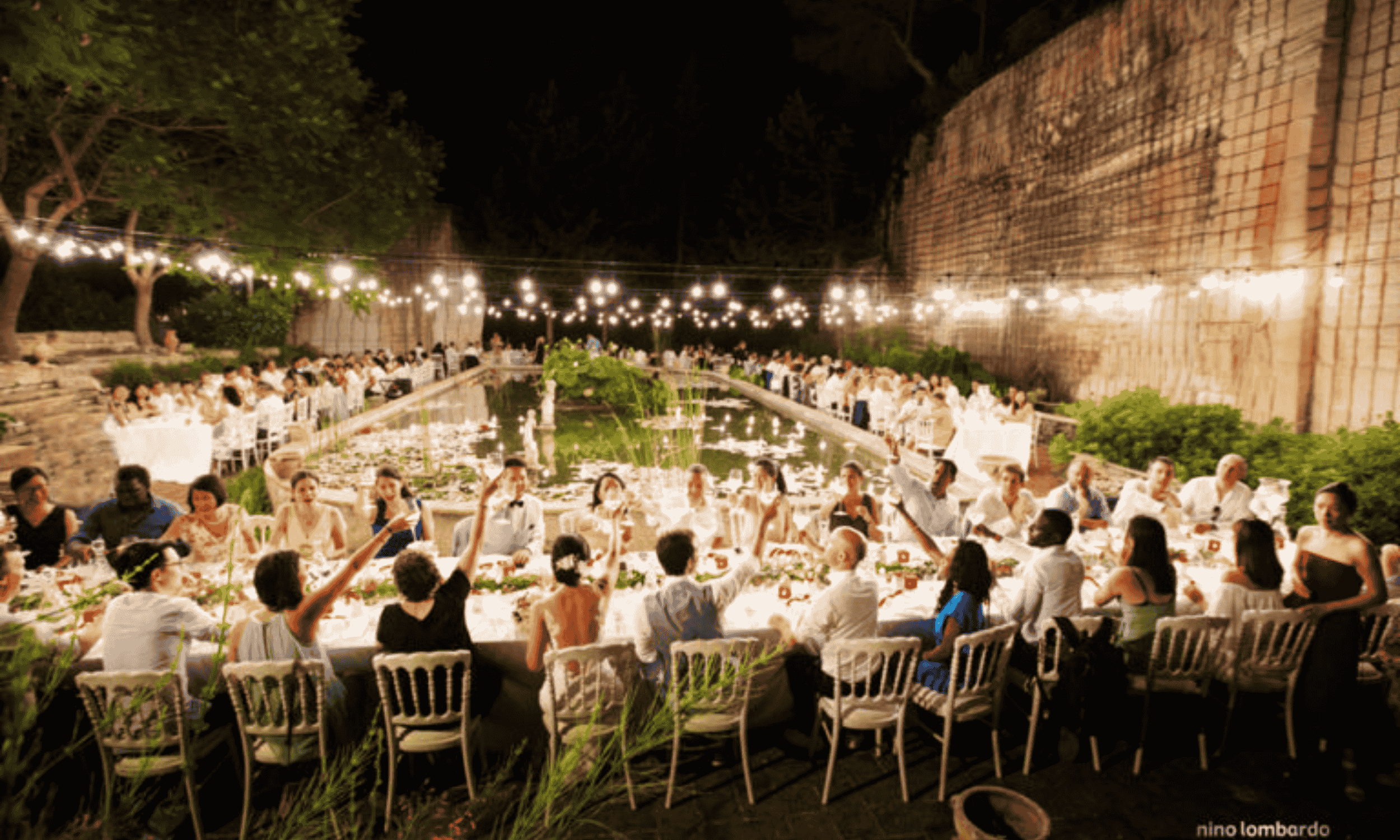 Guests seated at long outdoor wedding reception tables in Italy around a central water feature with string lights at night