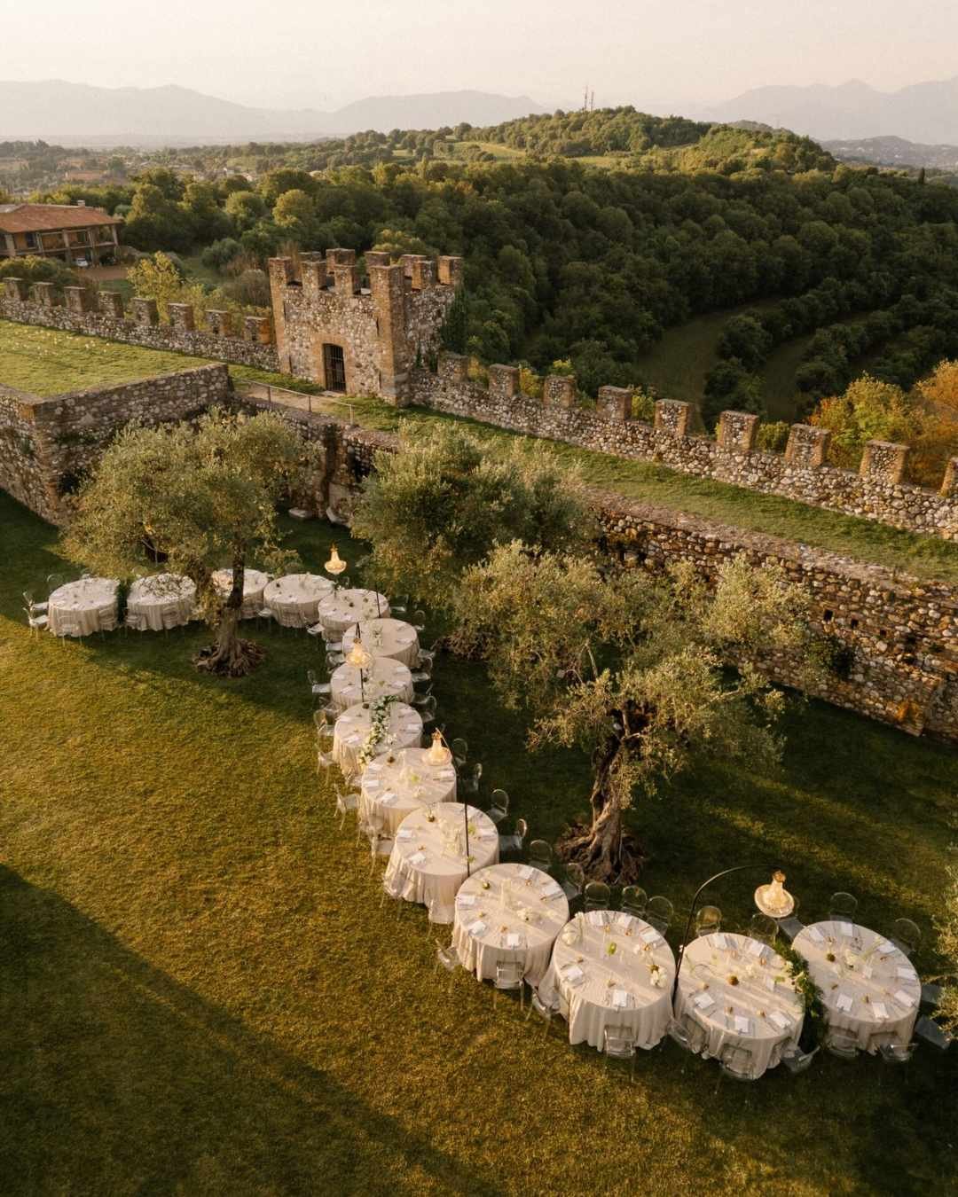 Aerial view of curved round tables flowing through olive trees beside an Italian castle, styled for a modern destination wedding.