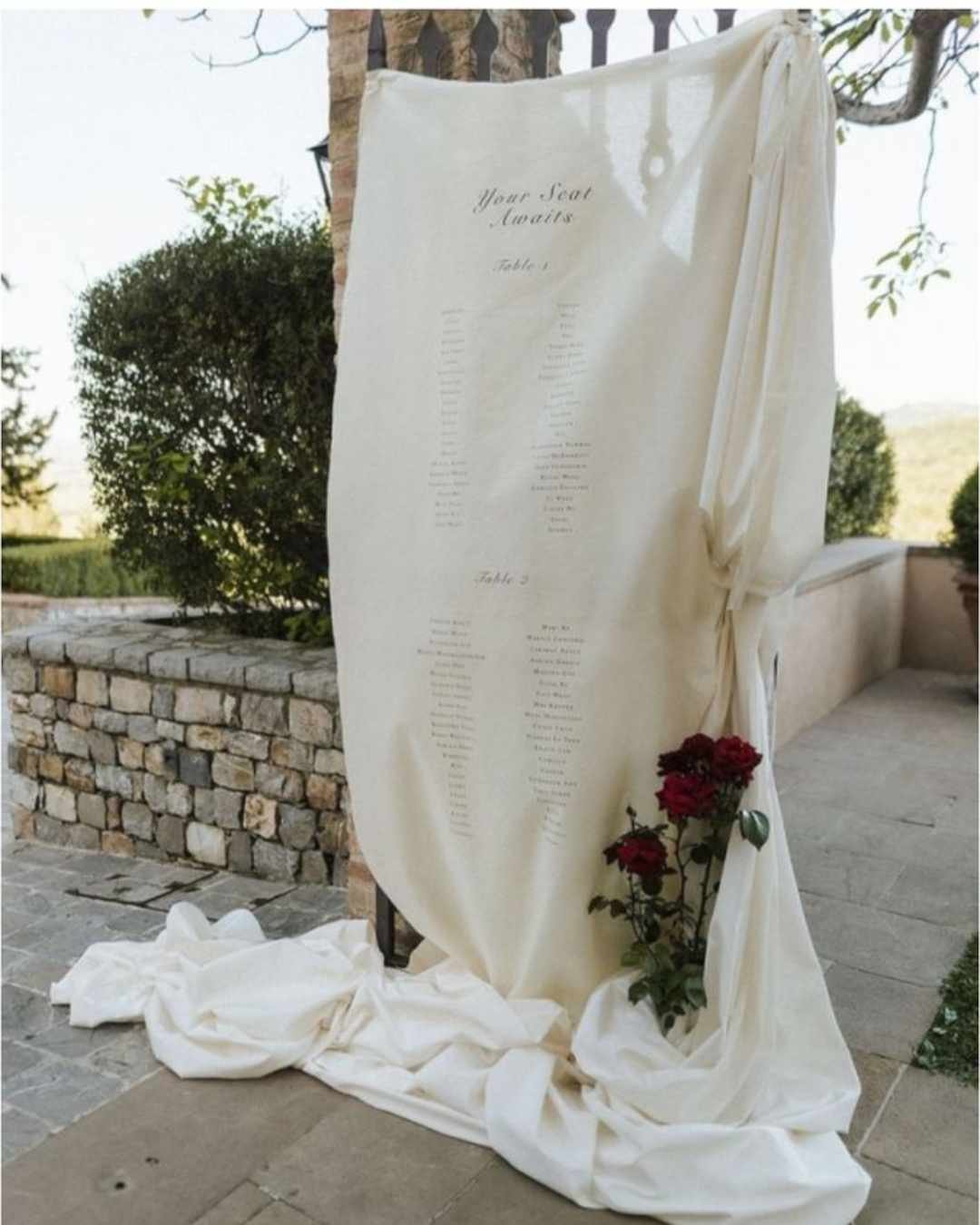 Draped fabric seating chart with red roses at an Italian wedding, blending minimal design with romantic textures.