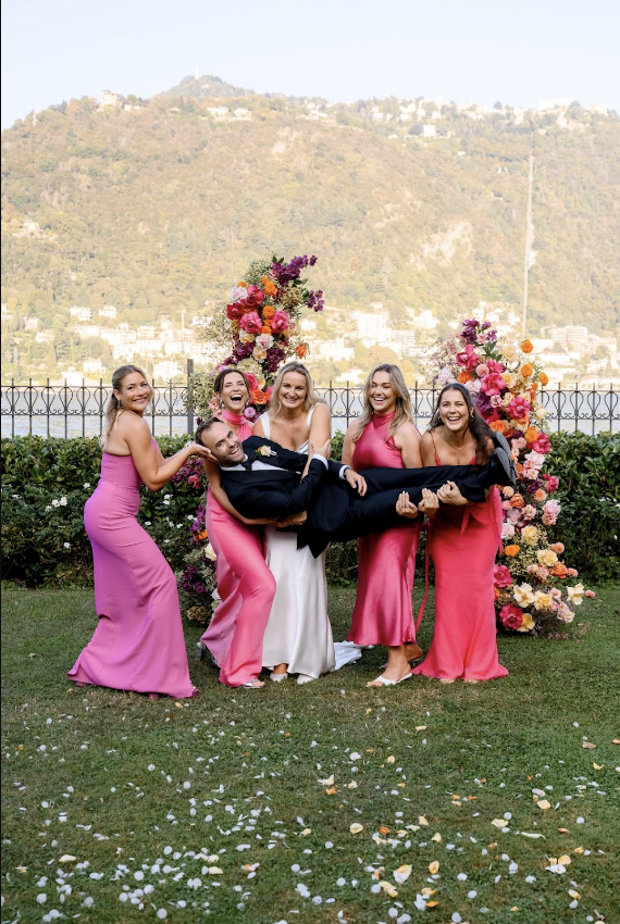Bride and bridesmaids laughing while holding the groom during a joyful outdoor wedding celebration.