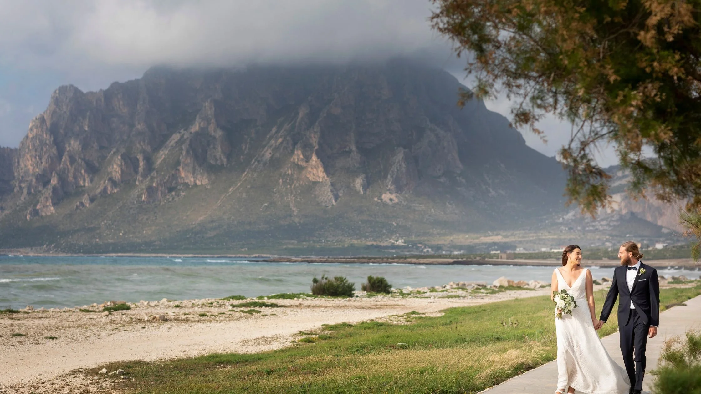 Bride and groom walking hand in hand along the Sicilian coast in Trapani, with dramatic mountains and the Mediterranean Sea as a backdrop.