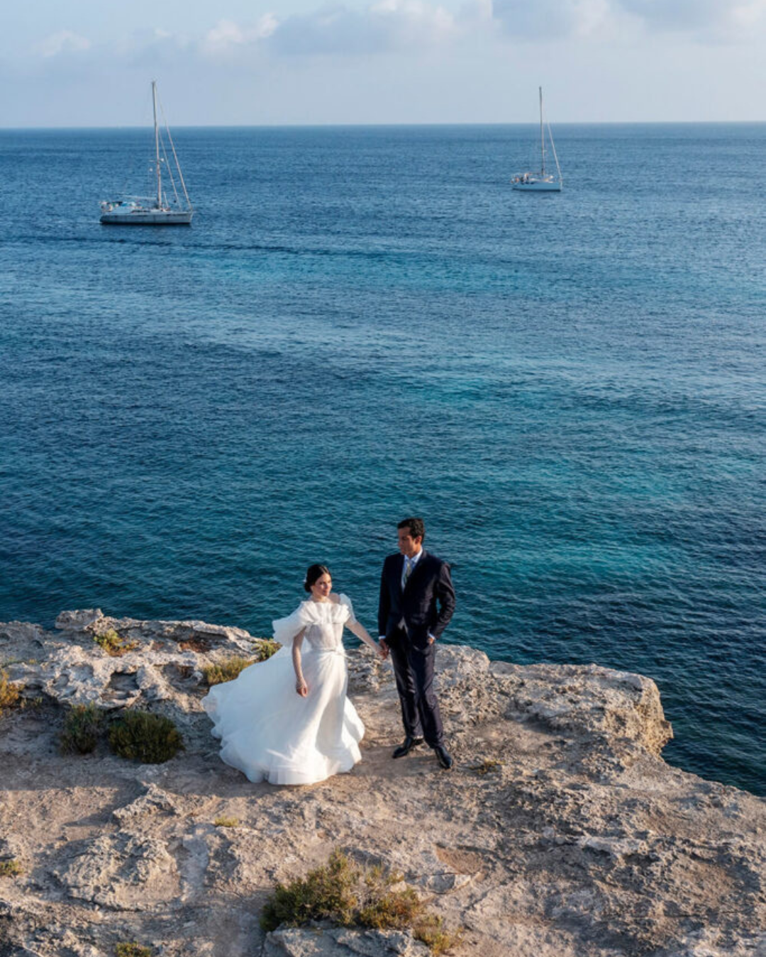 Bride and groom standing on a rocky cliff overlooking the sea in Italy with sailboats in the distance and a wide coastal view