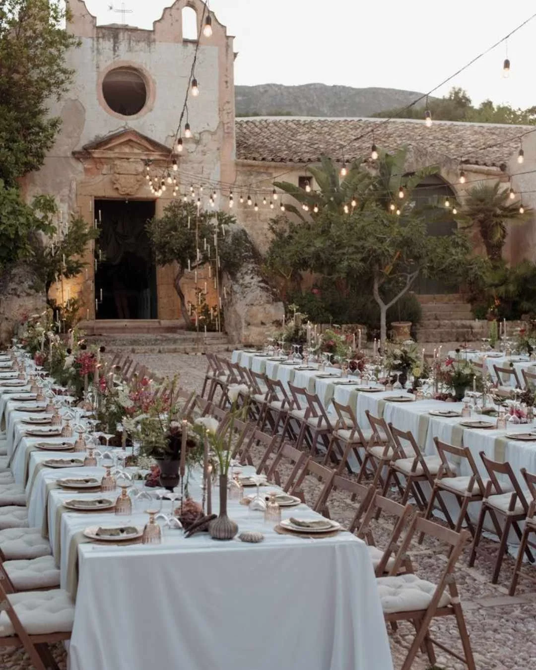 Long wedding tables styled with soft florals outside a rustic Italian church, set for an intimate 2026 destination wedding.