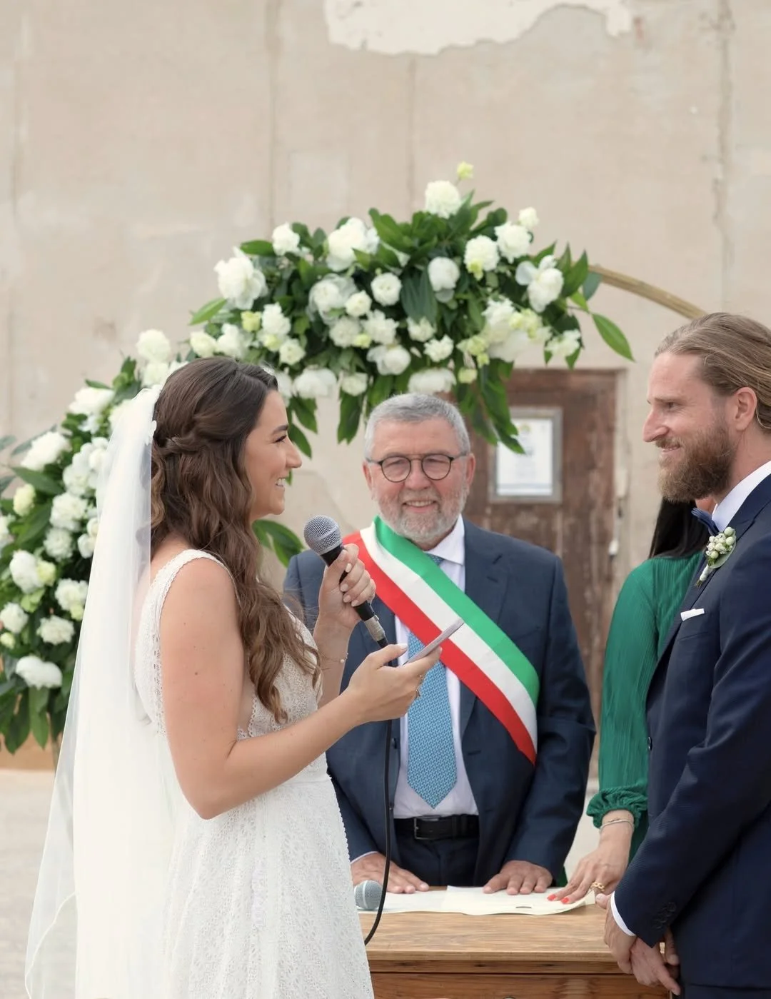 Bride speaking during an intimate wedding ceremony in Sicily, Italy, while holding a microphone beside local celebrants and guests.