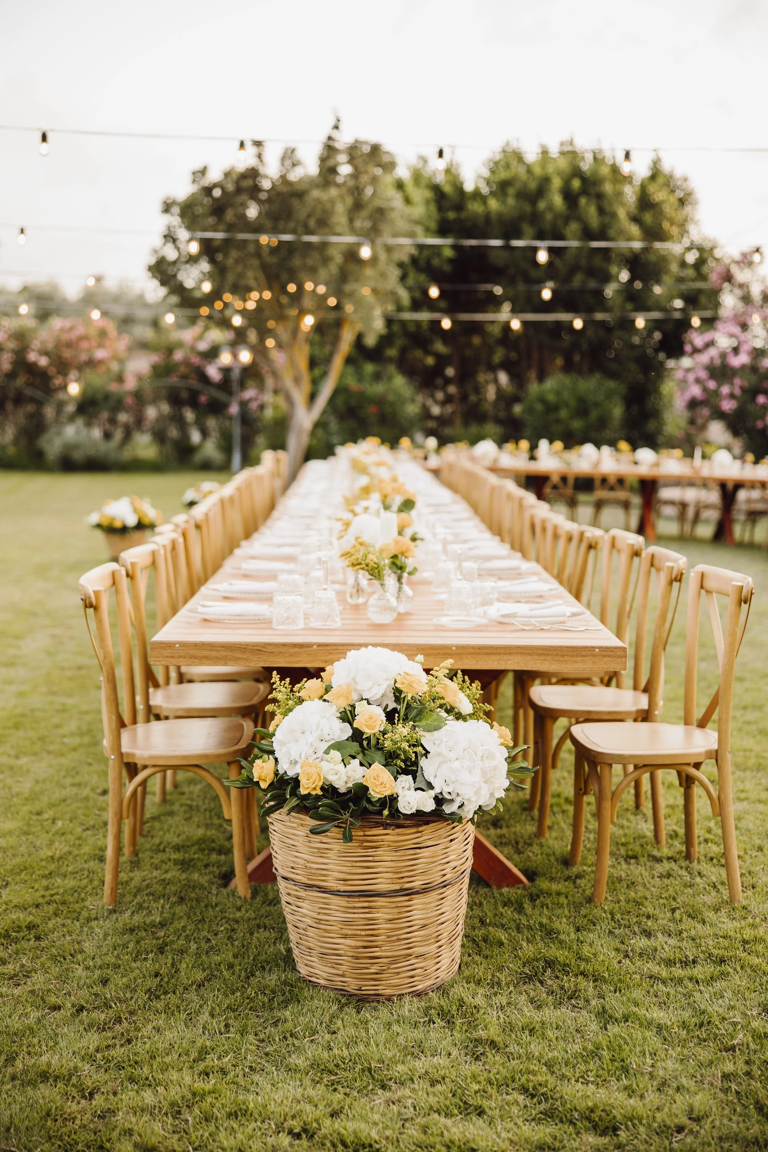 Garden wedding reception in Sicily with wooden tables, flower baskets, soft yellow and white florals, and romantic string lights by Alla Flora Giapponese.