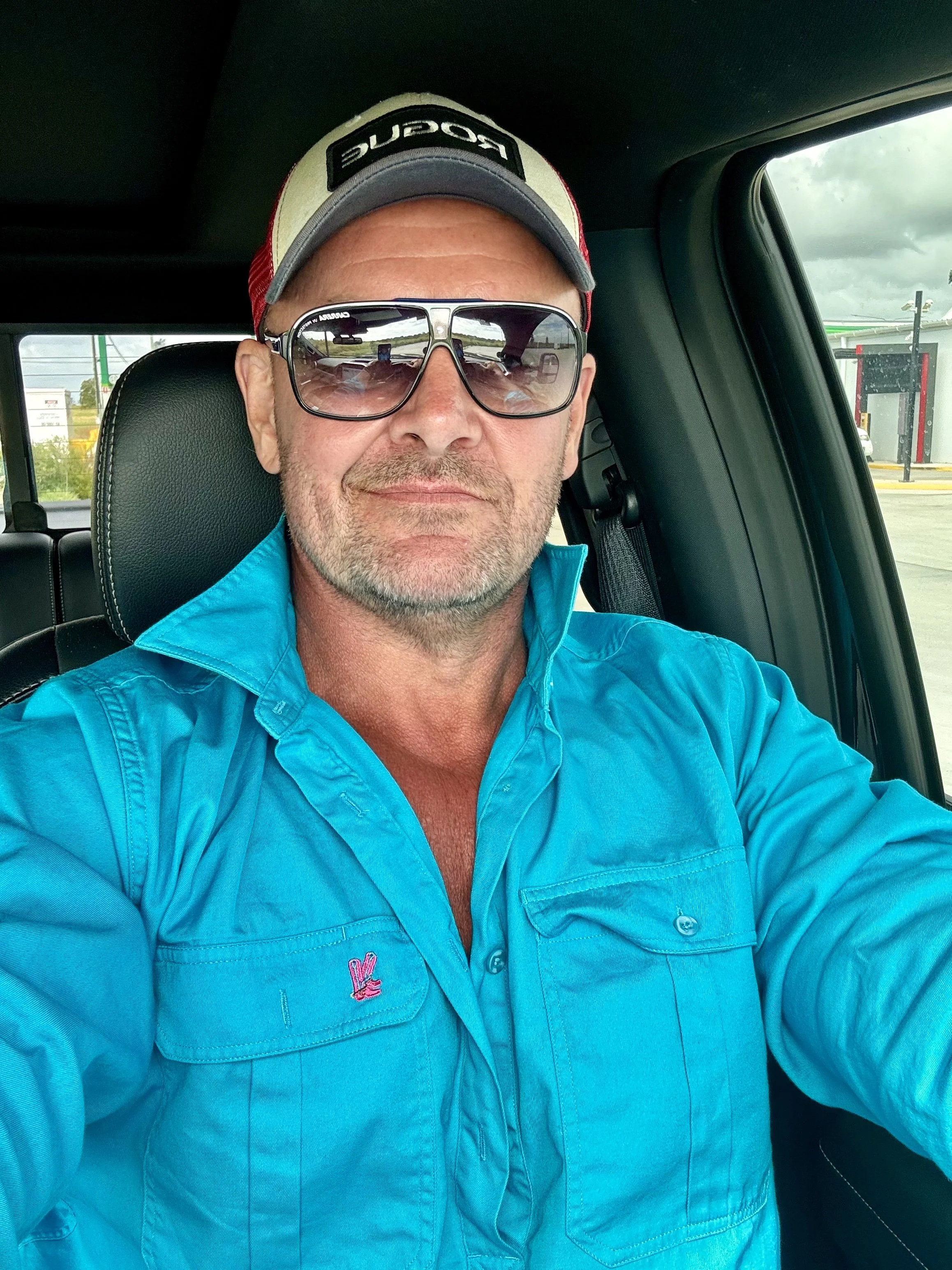 Man wearing sunglasses and a cap, seated inside a vehicle, dressed in a blue shirt with a visible pink logo.