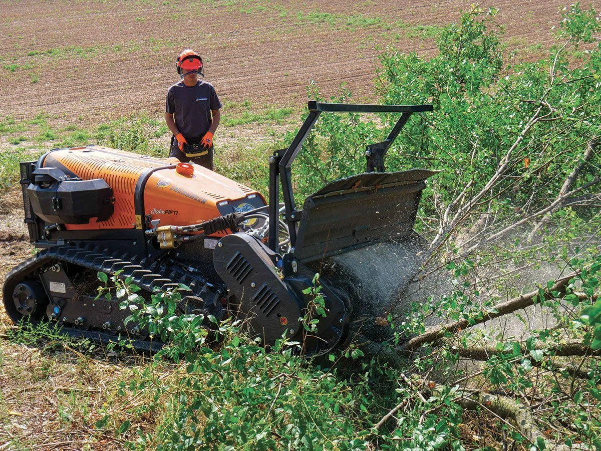 A person wearing protective gear using a remote-controlled orange forestry mulcher to clear vegetation in a field.