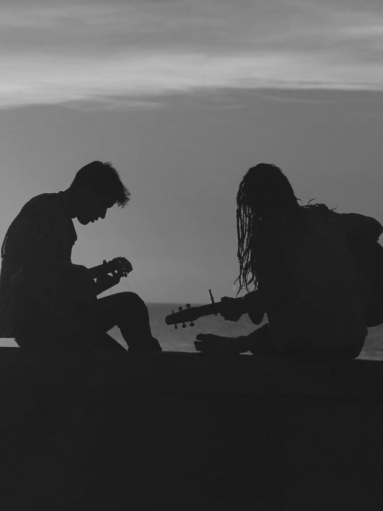 Silhouettes of two people sitting outdoors, one holding a guitar and the other holding a camera, during sunset or sunrise.