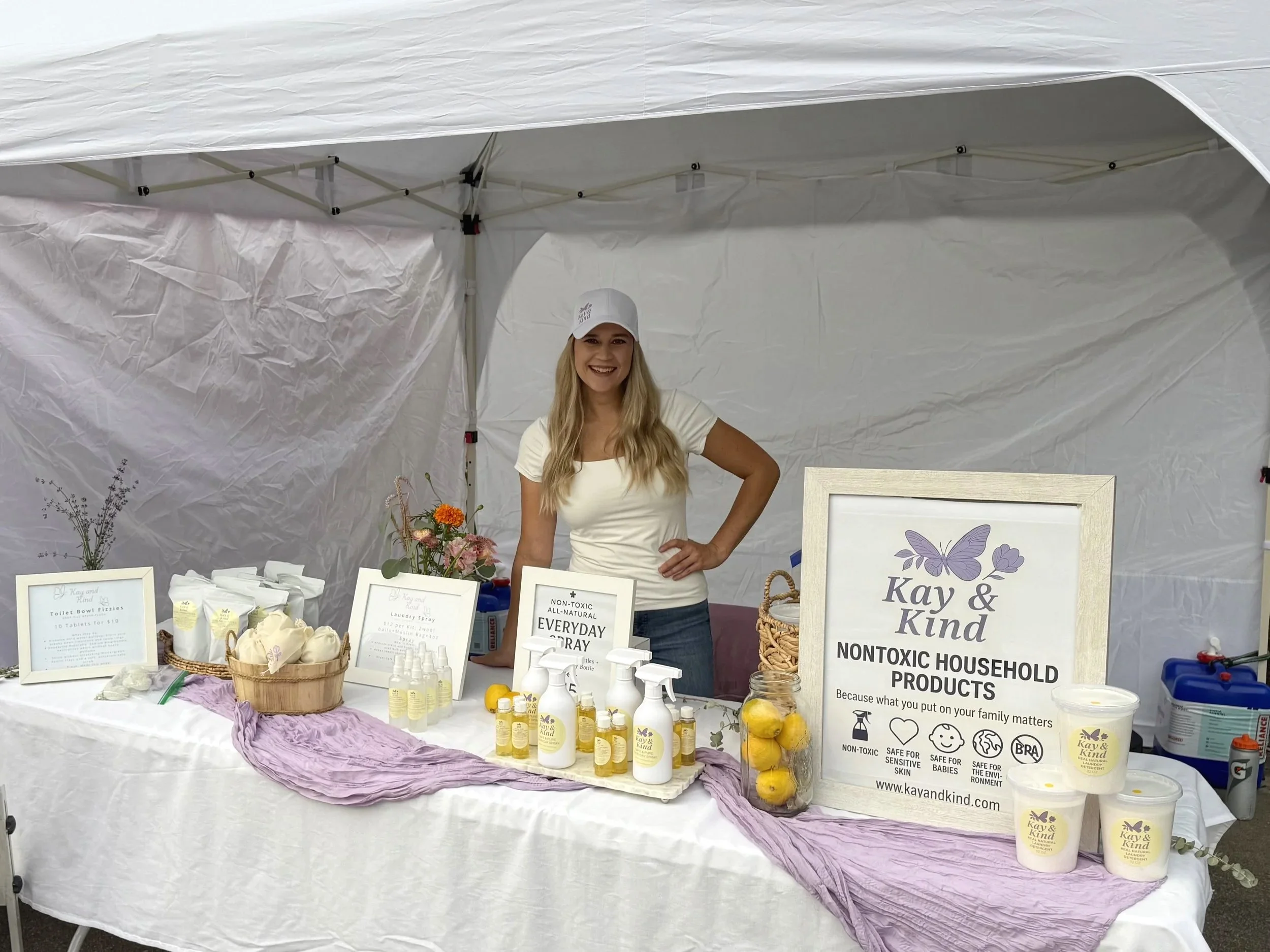 A woman standing behind a table of non-toxic household products at an outdoor market. The table has bottles, candles, and framed signs promoting eco-friendly products. The woman is smiling, wearing a white hat and a white shirt inside a white tent.