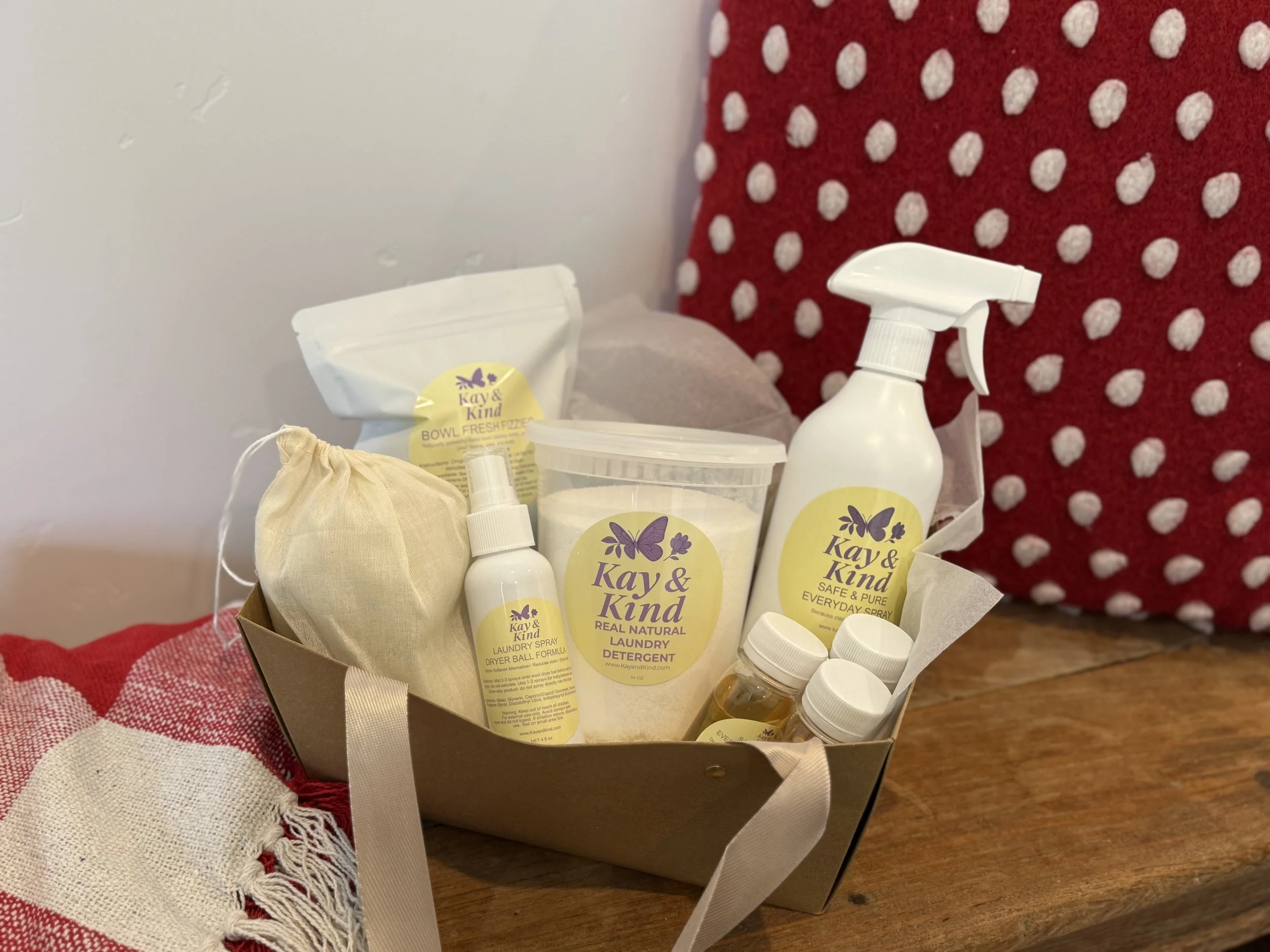 A gift basket containing eco-friendly laundry and cleaning products with yellow labels featuring purple butterflies, placed on a wooden surface with decorative textiles and a red and white polka-dot pillow in the background.