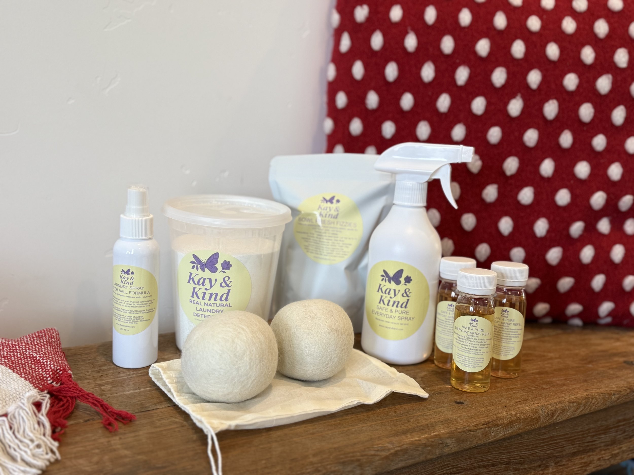 Collection of natural laundry and cleaning products, including sprays, powders, and refill bottles, arranged on a wooden surface with wool dryer balls and a red and white patterned pillow in the background.