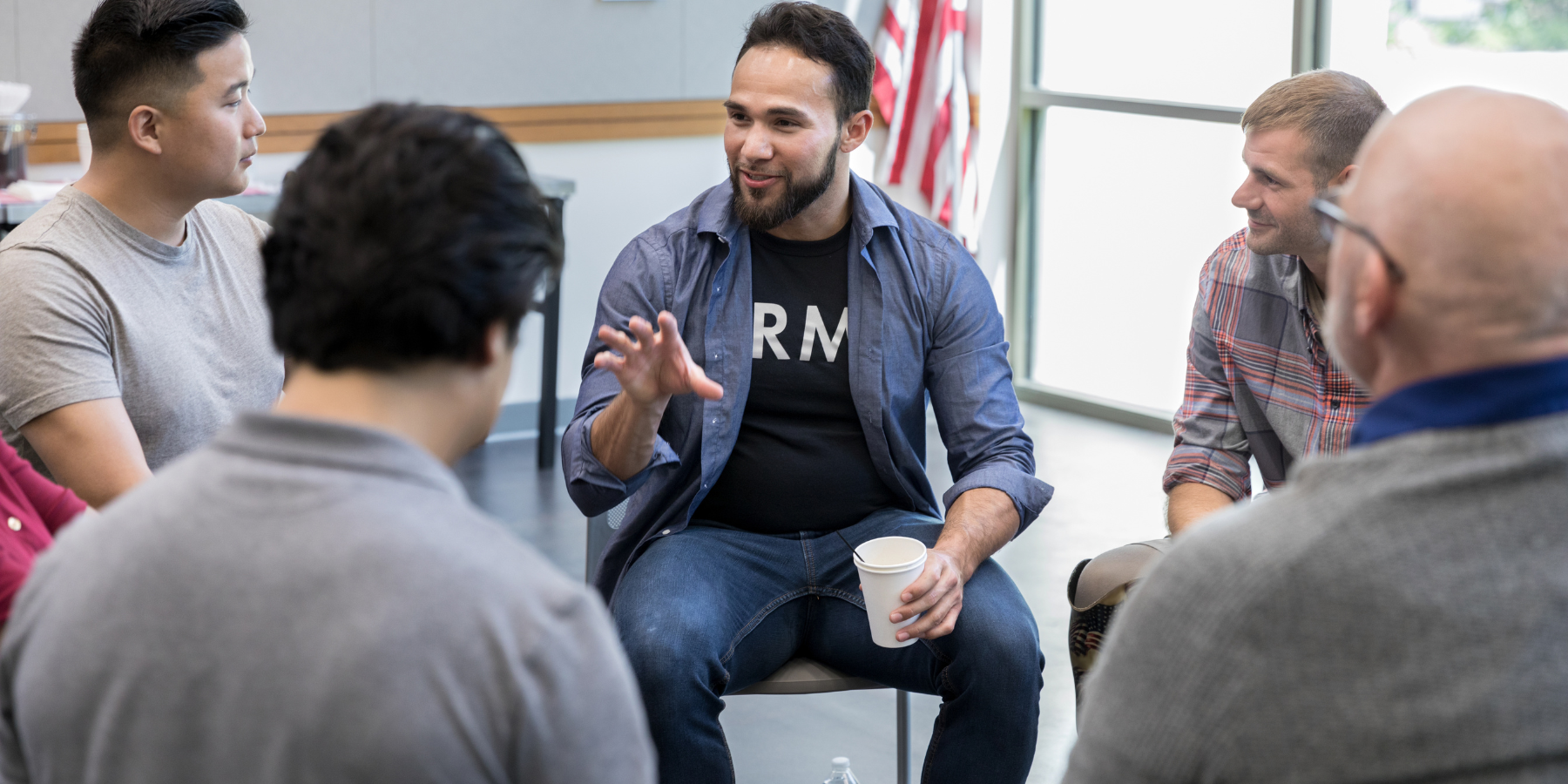 A diverse group of people sitting in a circle having a discussion. One man, holding a cup, is gesturing while talking. An American flag is visible in the background.