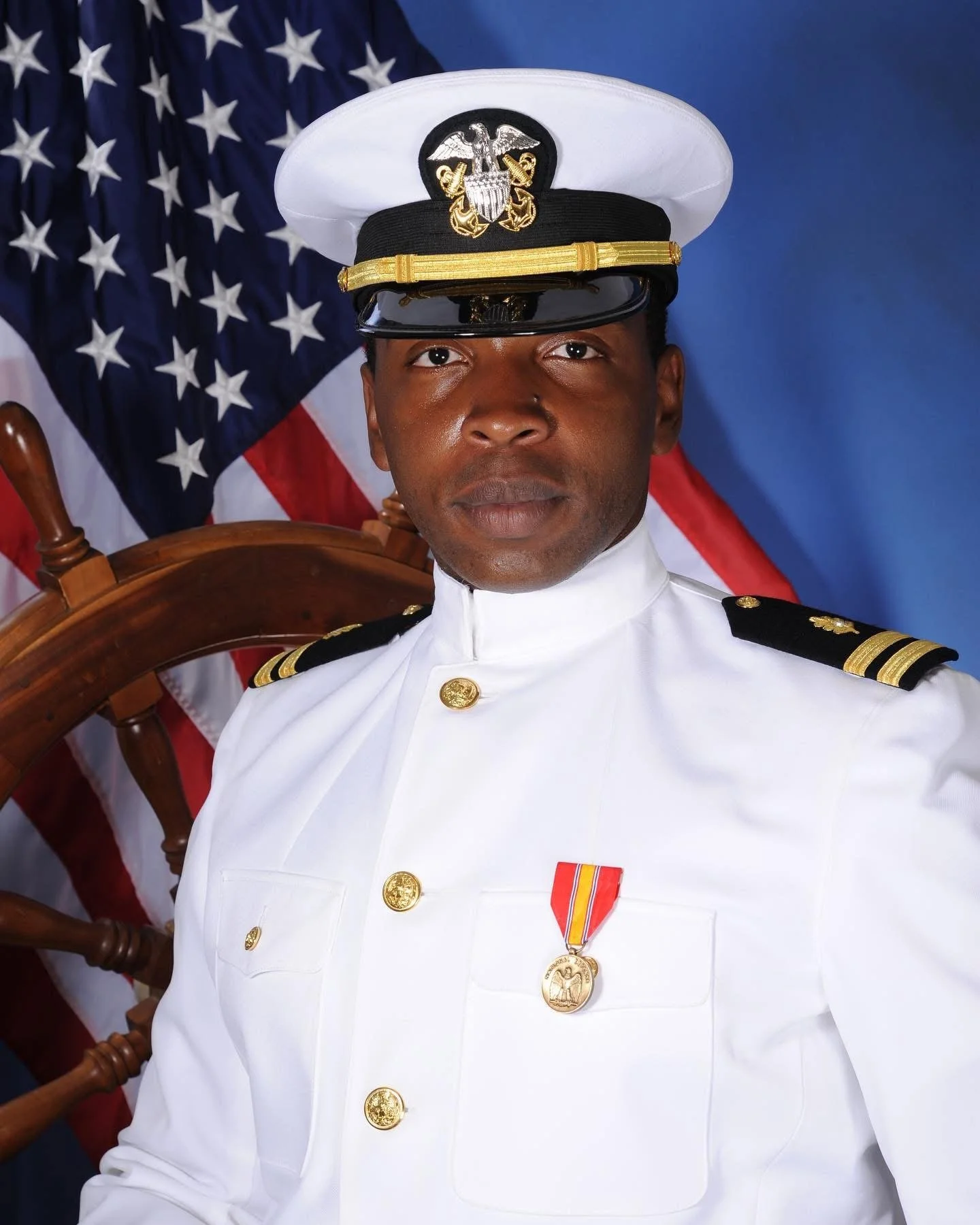 Navy officer in white uniform with a medal, in front of a U.S. flag and ship's wheel.