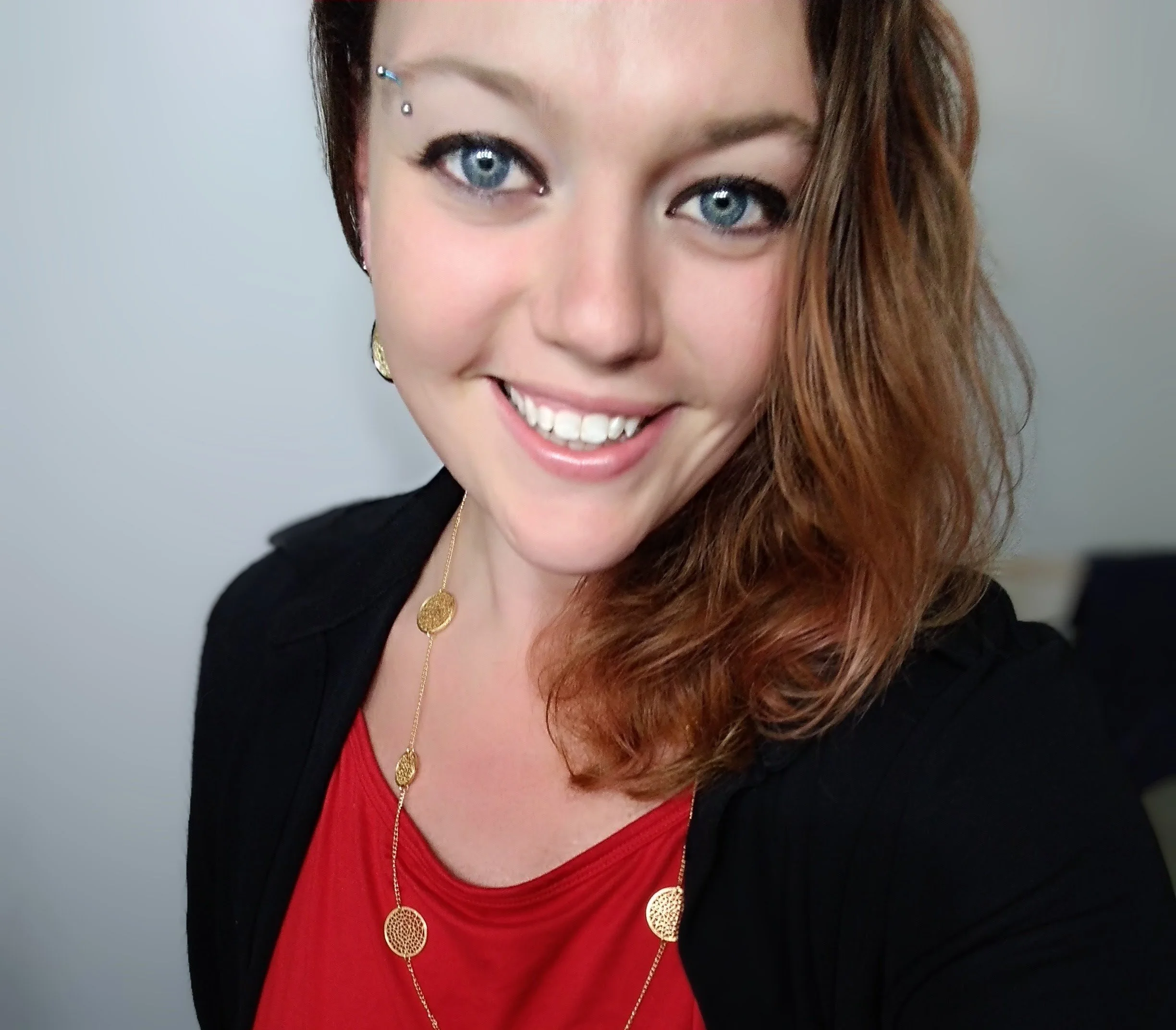 Woman smiling with blue eyes and brown hair, wearing a red top and gold necklace.